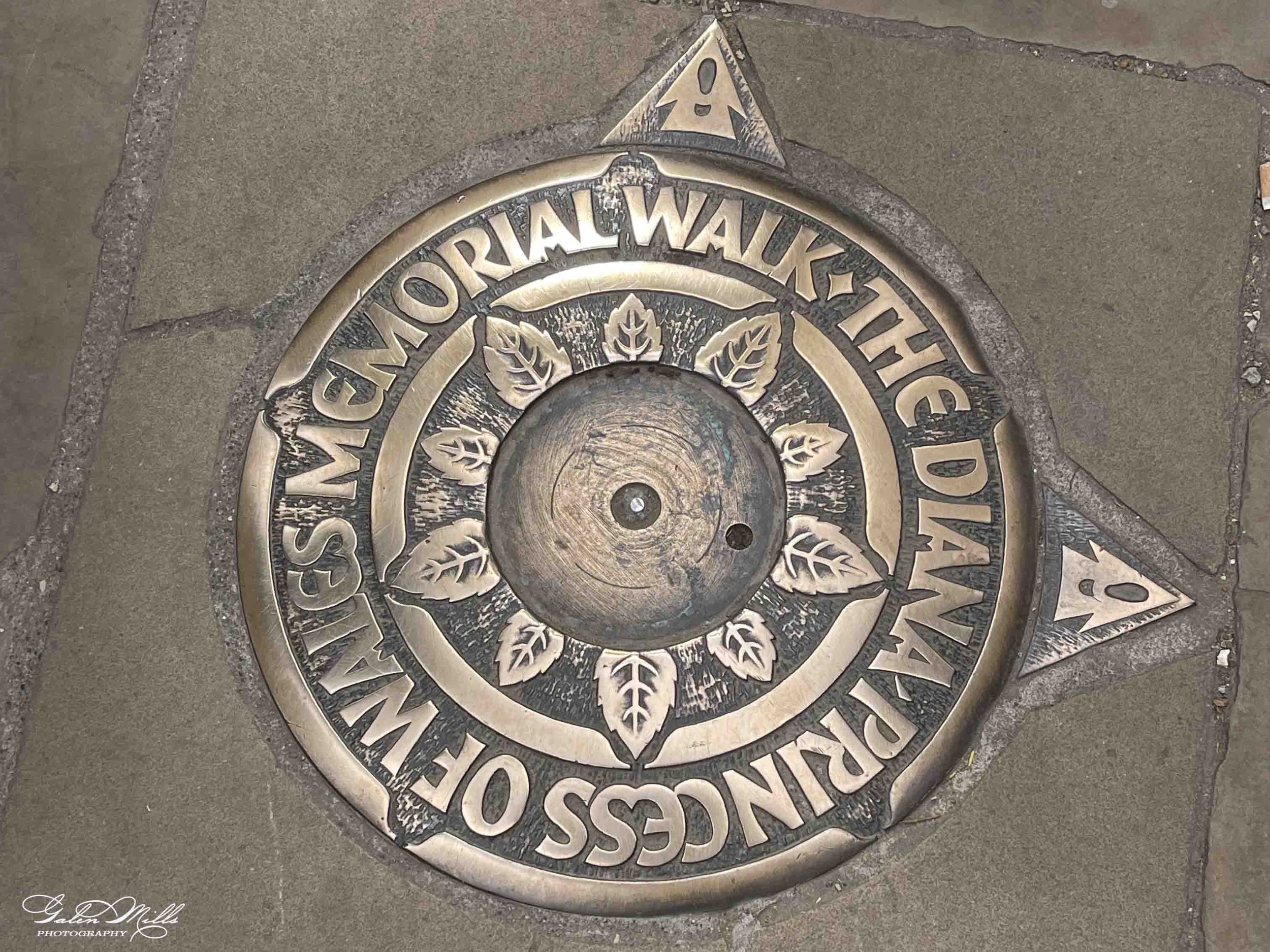 Close-up of a decorative sidewalk marker with the words 'NATIONAL WARDEN'S OF SCOTLAND' and an emblem with thistles and Scottish motifs, embedded in concrete pavement.