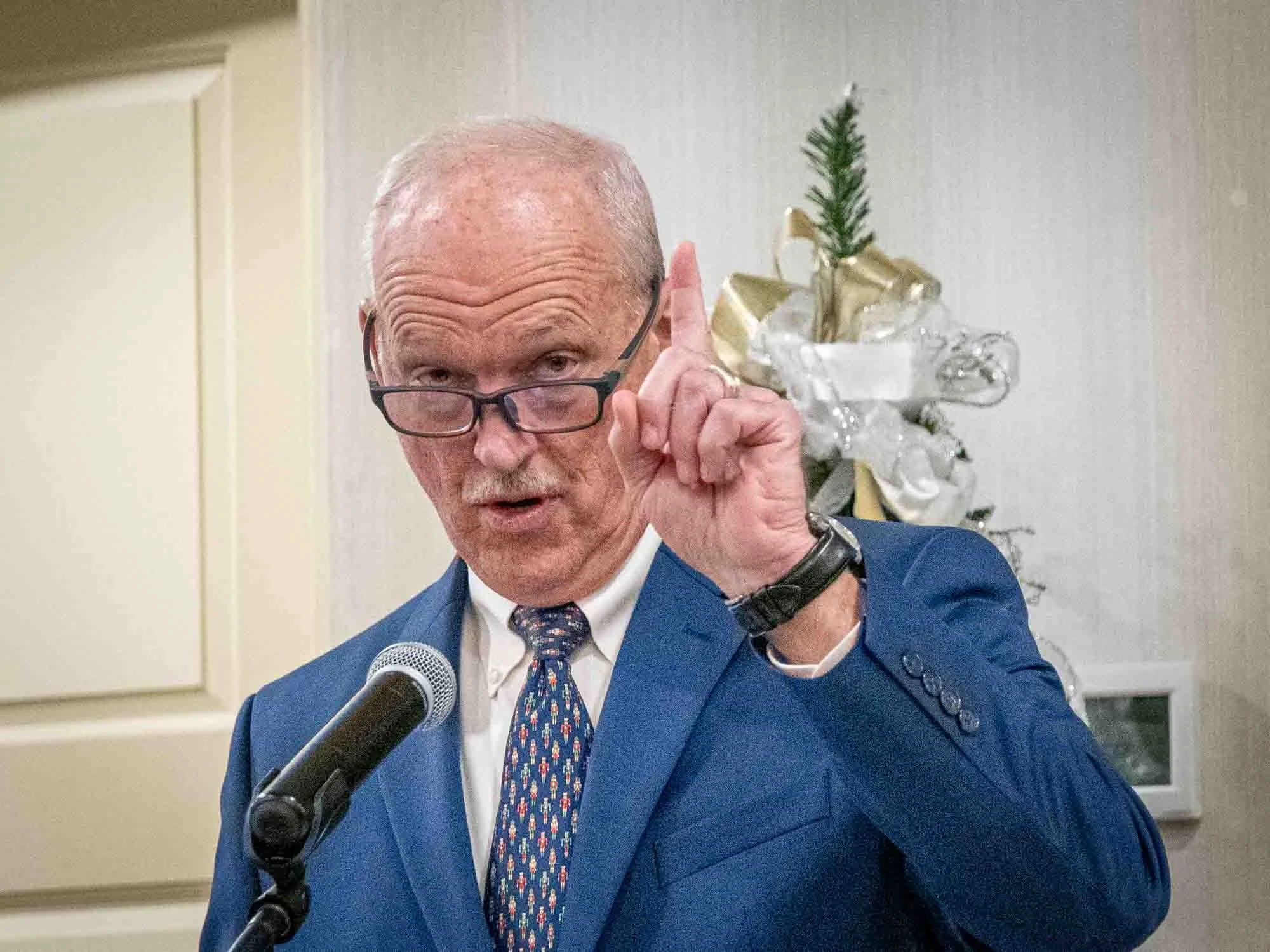 A man in a blue suit speaking into a microphone, gesturing with one finger raised, possibly making a point. He is wearing eyeglasses and a patterned tie. In the background, there is a decorative arrangement with ribbons and greenery.