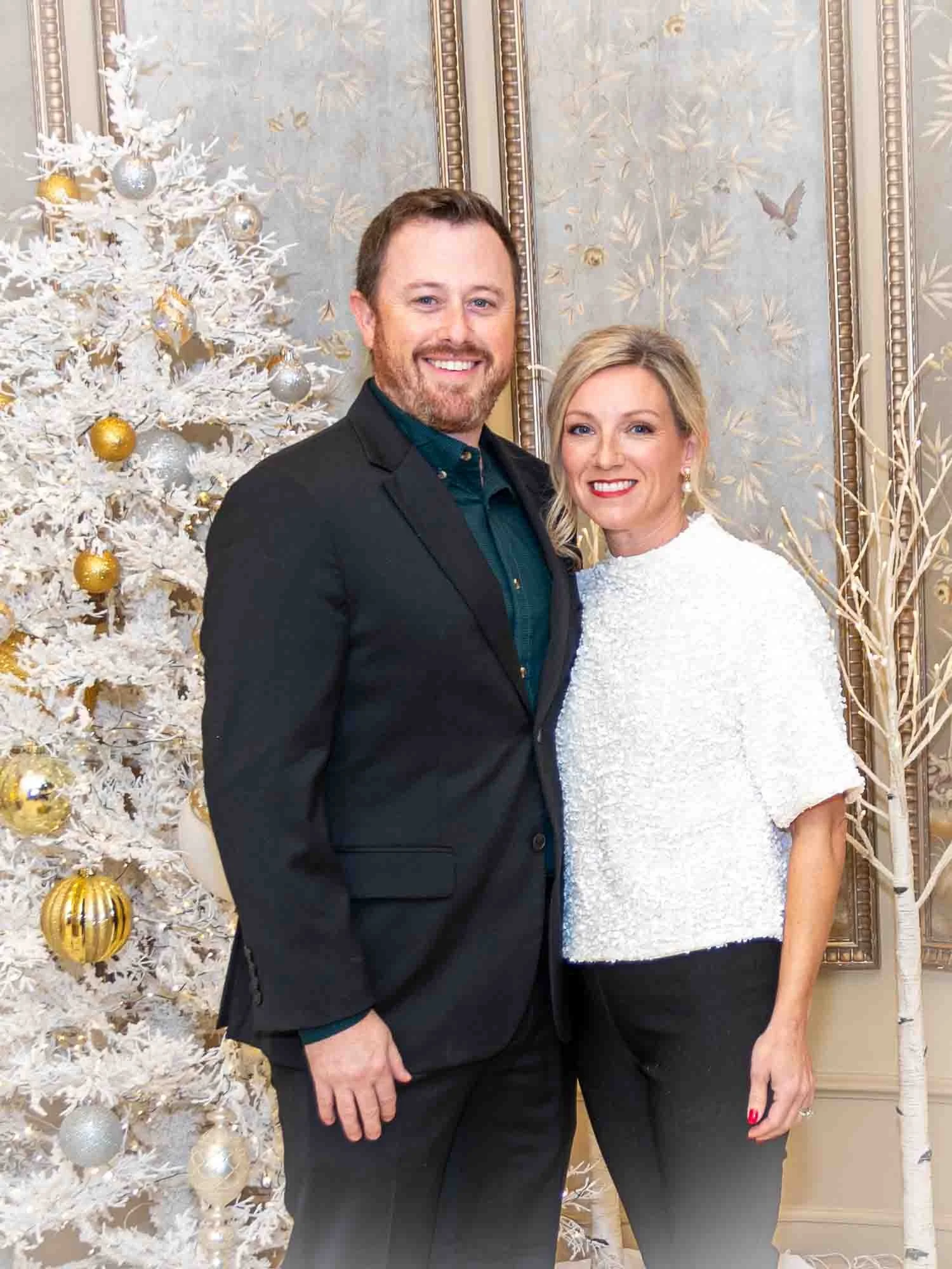 A man in a black suit and green shirt and a woman in a white blouse and black pants are standing in front of a decorated white Christmas tree with gold and silver ornaments, in an elegantly decorated room.