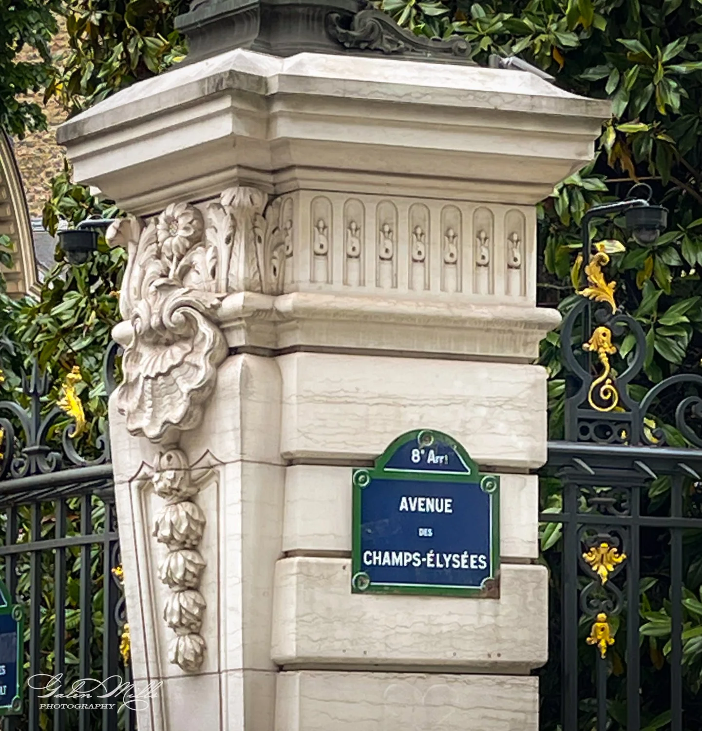 Street sign for Avenue des Champs-Élysées on a decorative stone pillar with ornate detailing and nearby ironwork gate.
