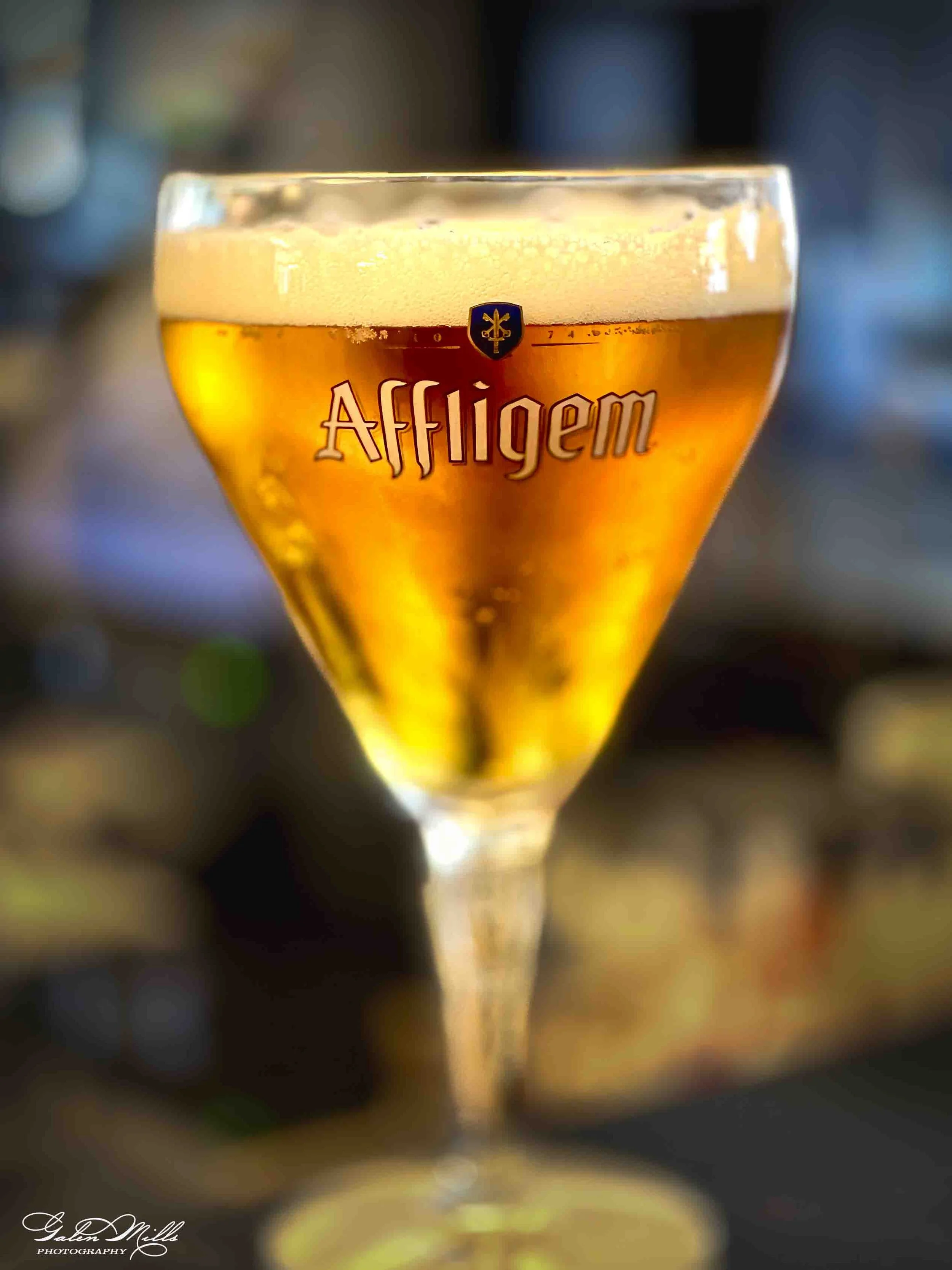Close-up of a glass of beer with a foamy head, the glass has the brand name 'Affligem' printed on it, and features a small shield crest logo above the brand name.