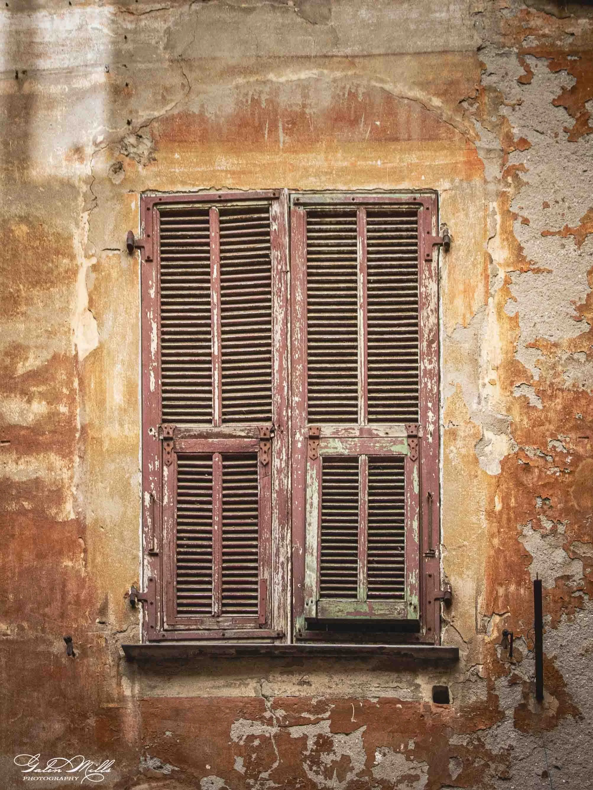 Old, weathered window with wooden shutters on a peeling, rustic wall.