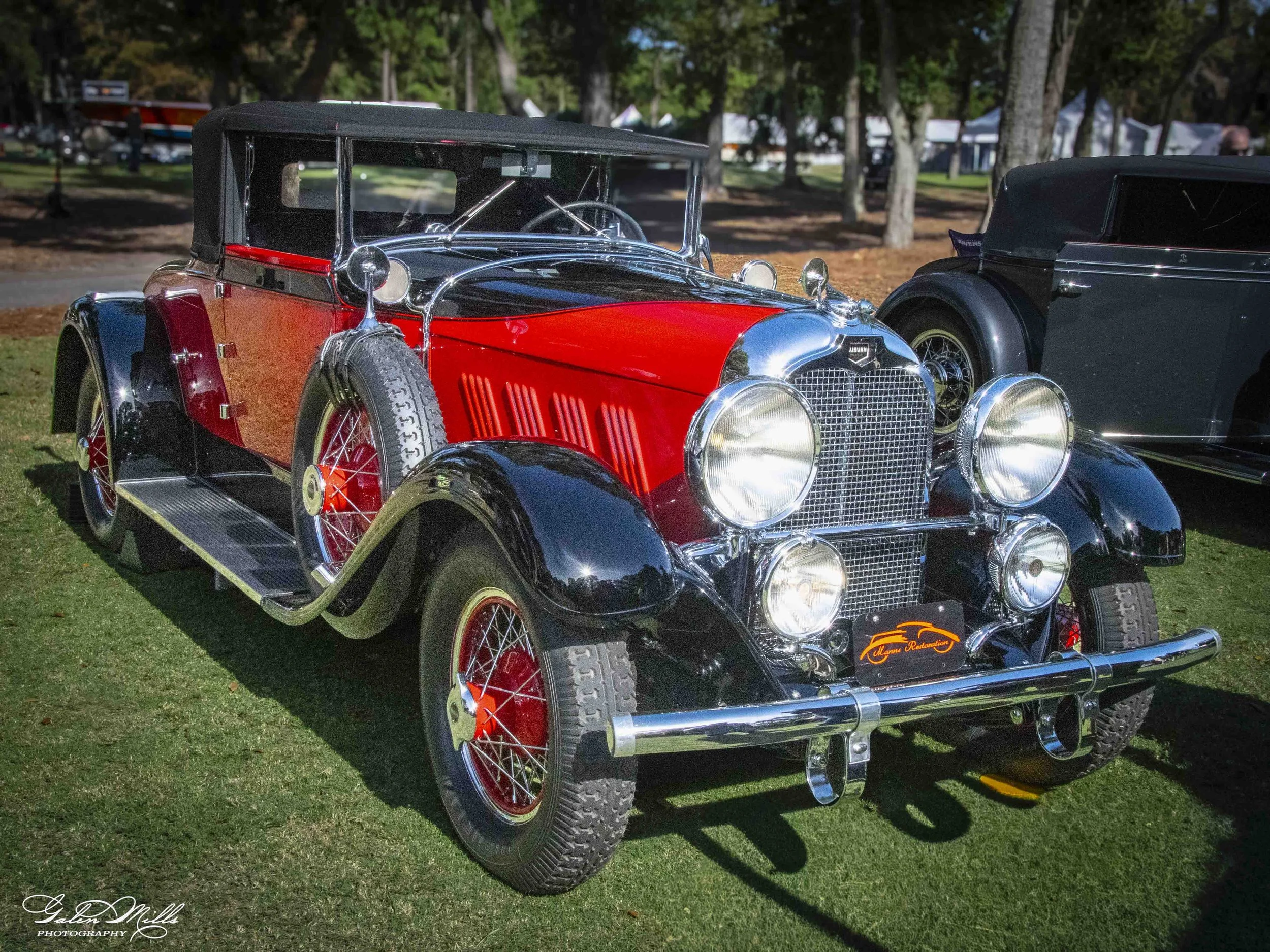 A vintage black and red automobile displayed outdoors on a grassy area at a car show or exhibition, with trees and tents in the background.