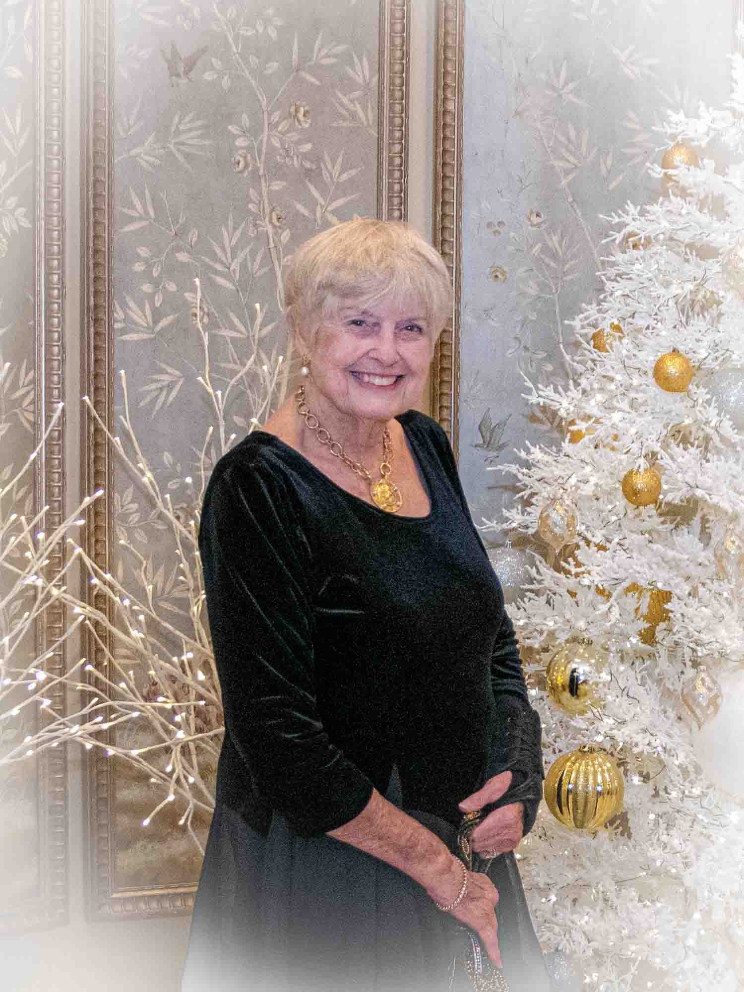 Elderly woman in black dress standing beside a white Christmas tree with gold ornaments.