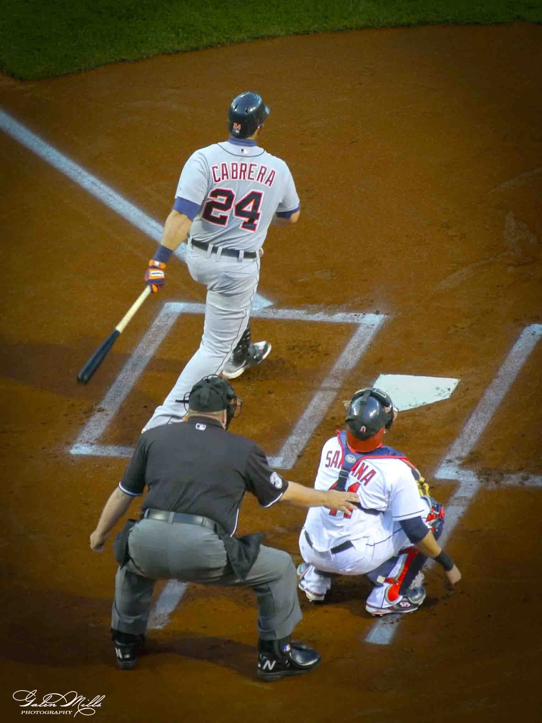 A baseball player in a gray uniform and black helmet runs towards home plate, while a catcher in a white and red uniform crouches behind the plate. An umpire in a black uniform approaches from the left.
