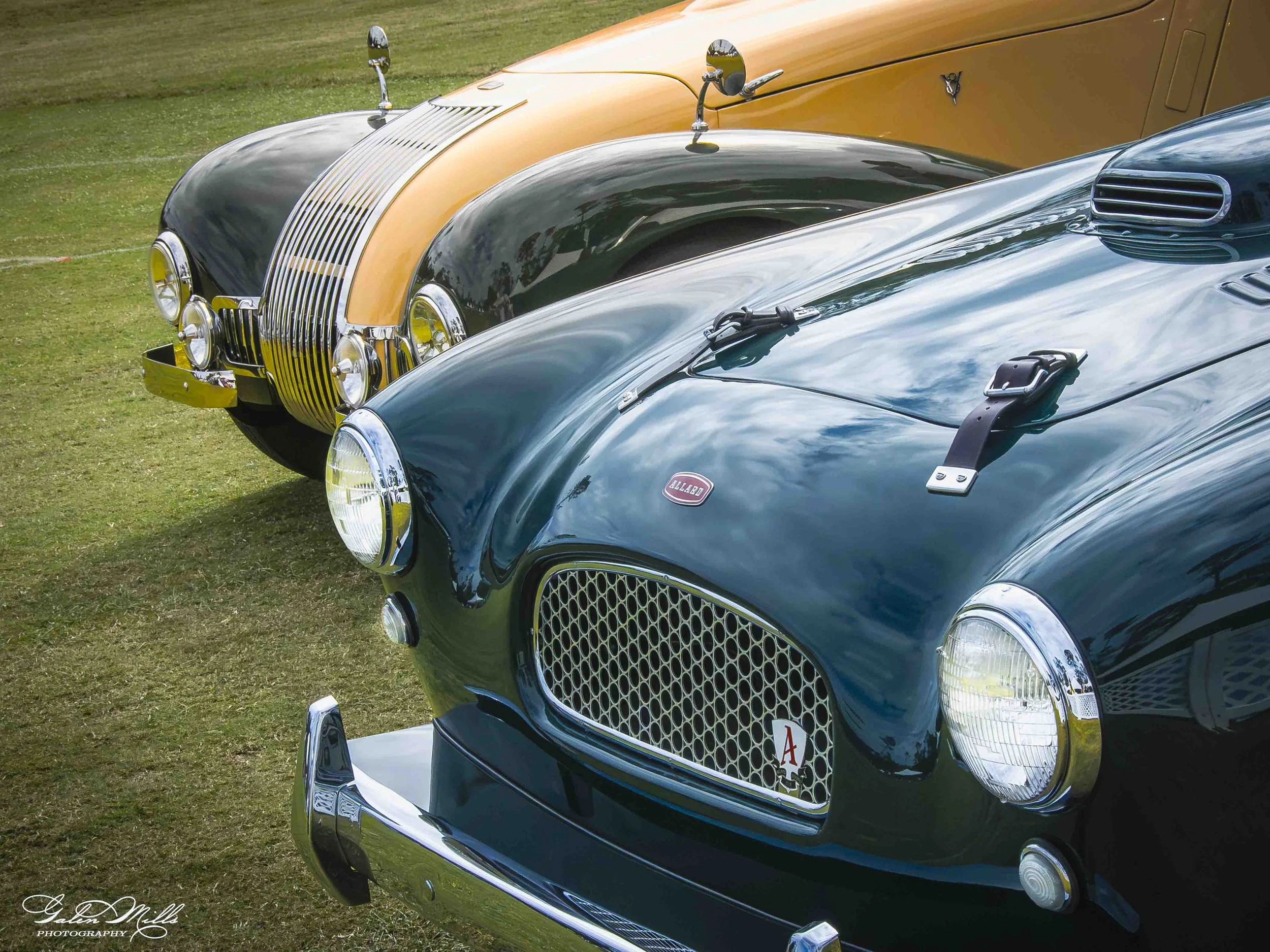 Close-up of the front ends of two vintage cars parked on grass, showcasing their chrome grilles, rounded headlights, and shiny paint in black and blue colors.