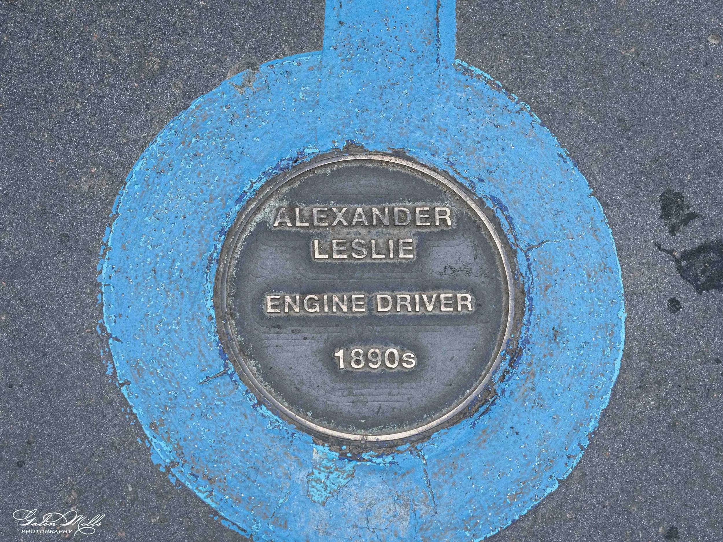 A manhole cover embedded in asphalt, marked with the name Alexander Leslie, indicating an engineer driver from the 1890s, surrounded by a blue painted circle on the pavement.