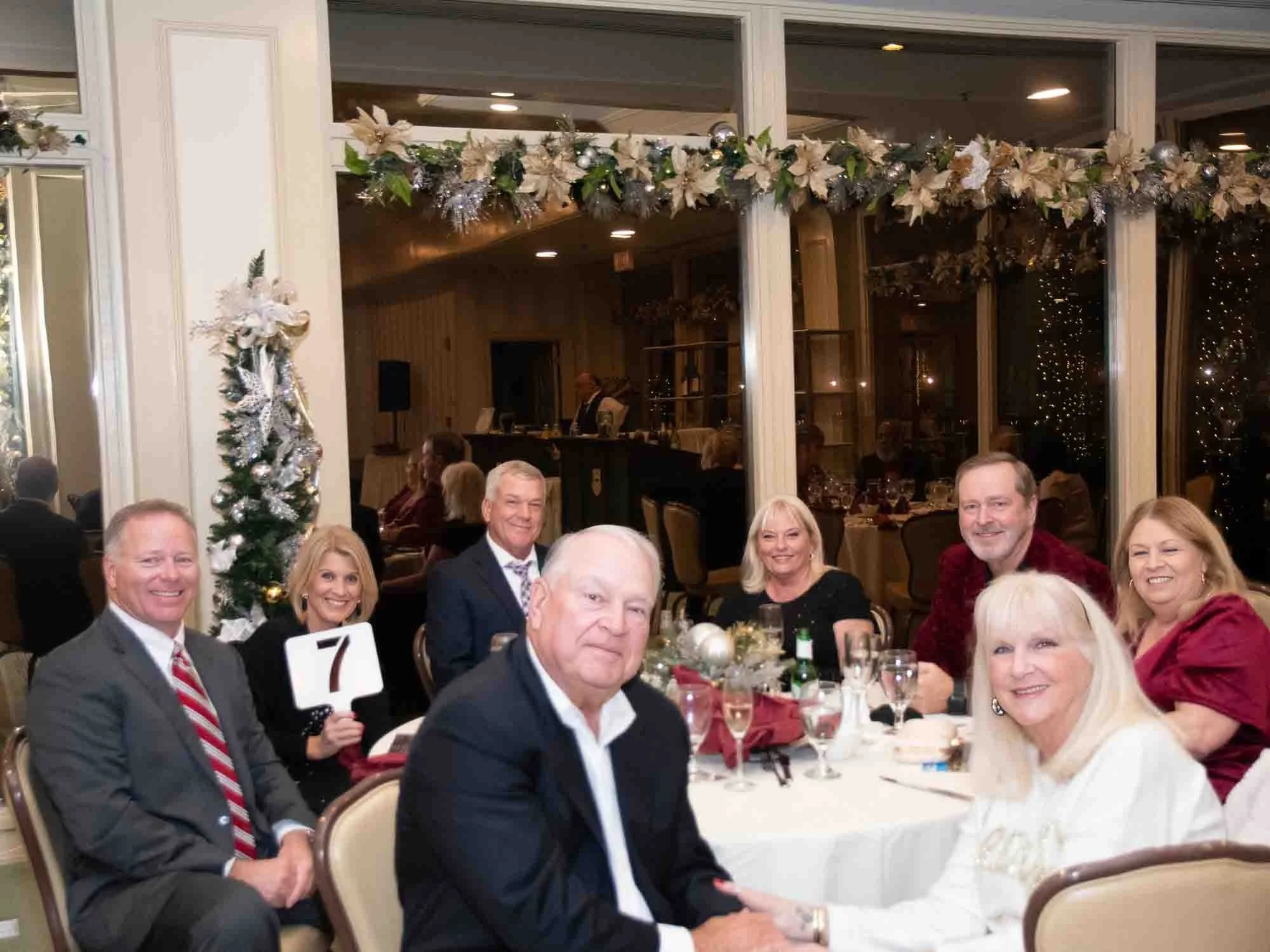 Group of people dressed formally sitting at a round table during a holiday party, with festive decorations and a small Christmas tree in the background.