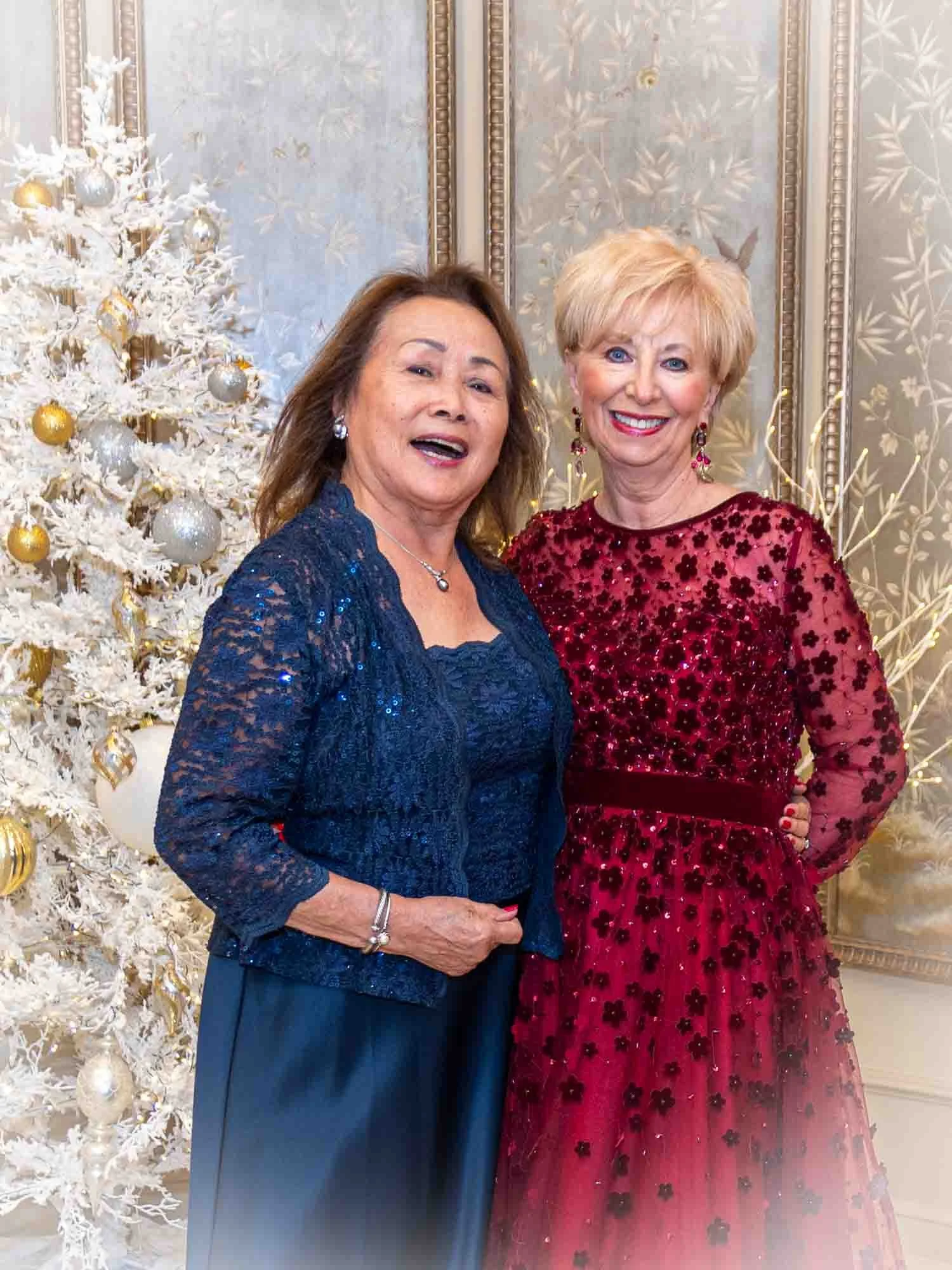 Two women dressed elegantly, one in a dark blue gown and the other in a red dress, standing in front of a decorative white Christmas tree with gold and silver ornaments.