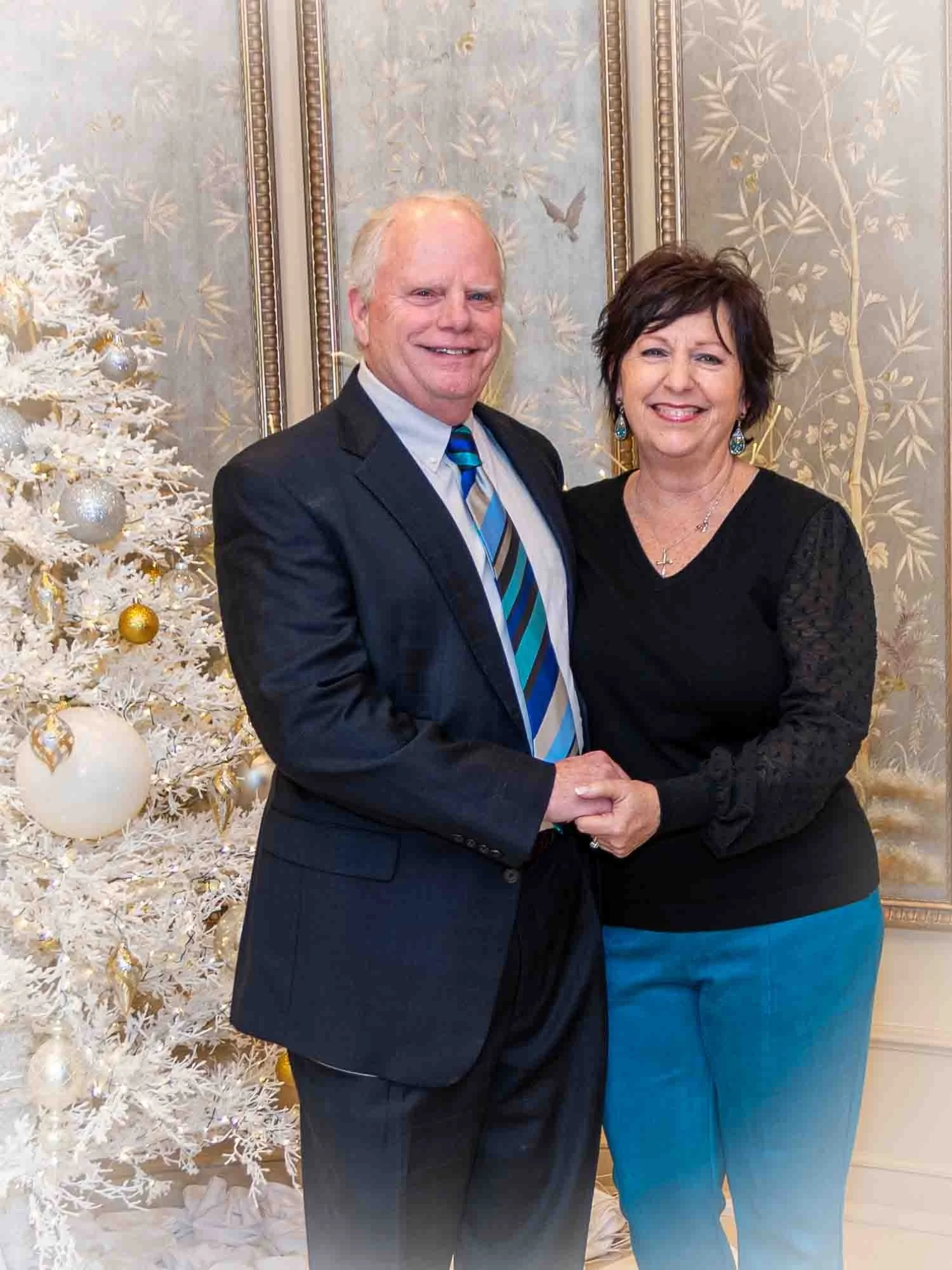 A couple smiling and standing close together indoors, with a decorated white Christmas tree in the background. The man is wearing a dark suit with a striped tie, and the woman is wearing a black top and blue pants. The setting appears festive.