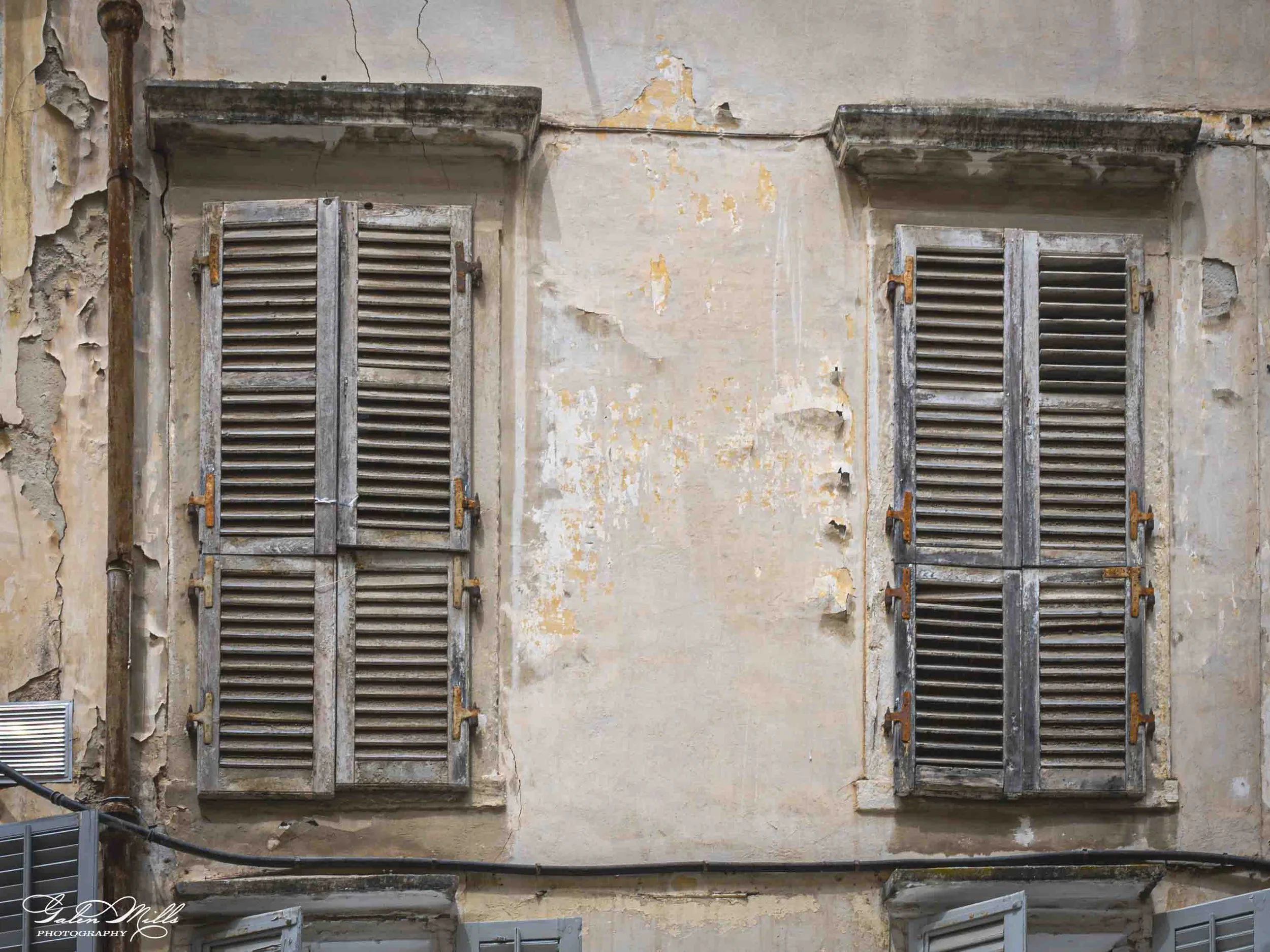 Four old wooden window shutters with louvered slats, mounted on a weathered, peeling wall.
