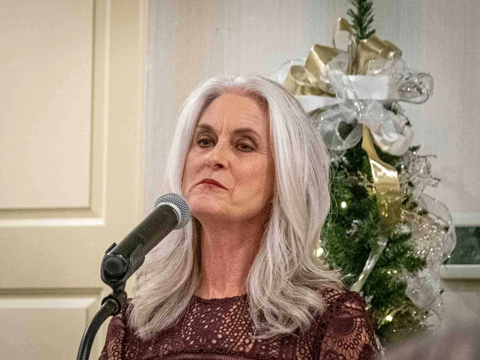 Woman with long gray hair speaking into microphone in front of decorated Christmas tree.