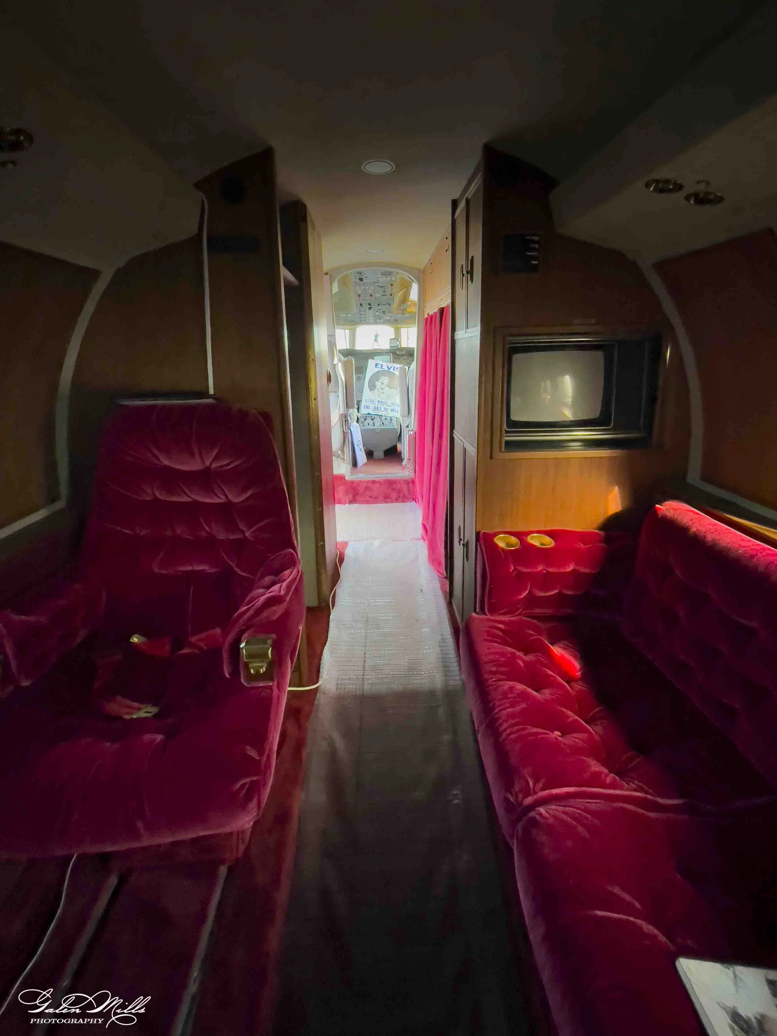 Interior of a vintage airplane with red velvet seats, a small TV, and a narrow aisle leading to the cockpit.