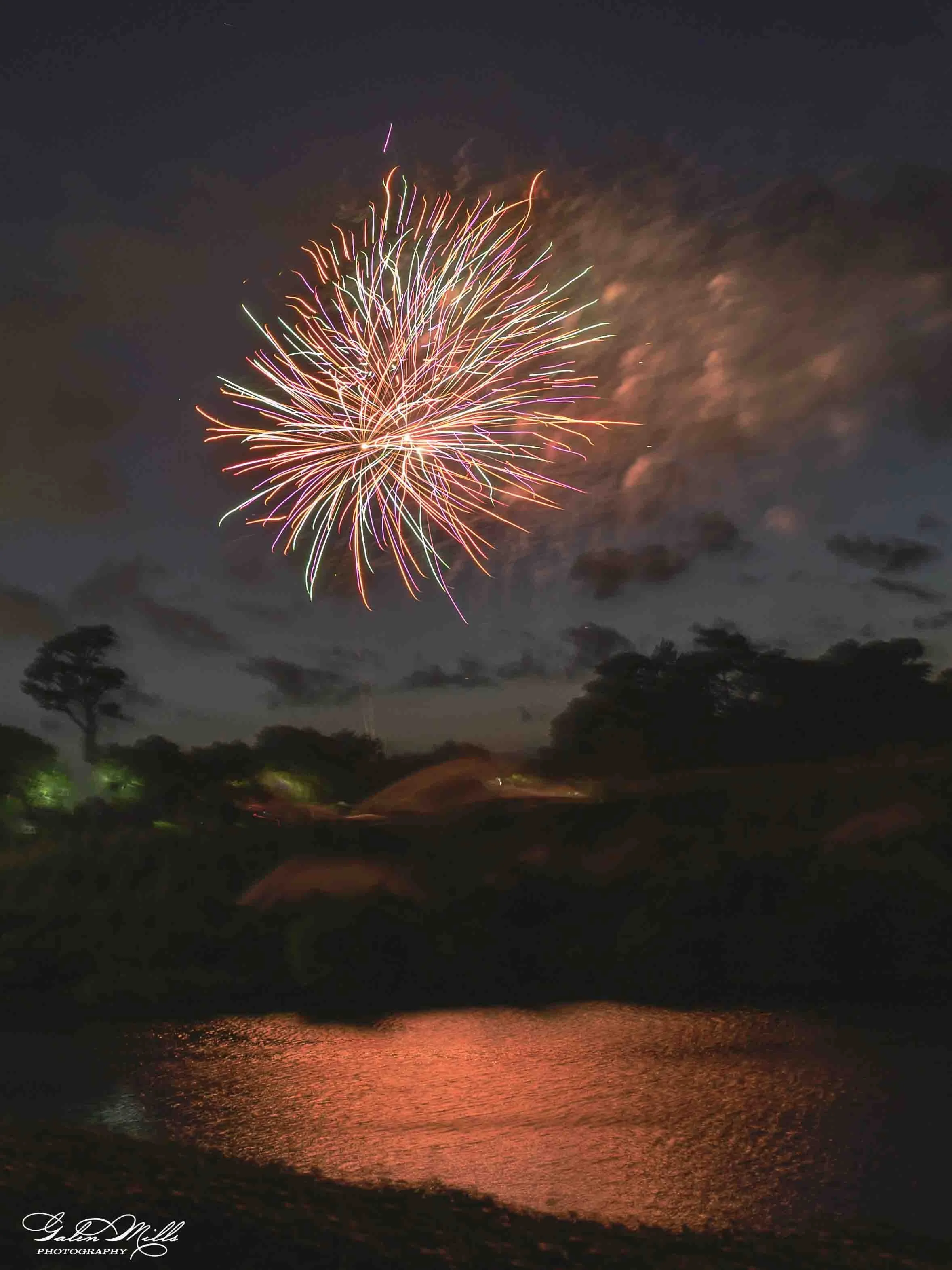 Nighttime fireworks over a calm body of water, with reflections on the surface and silhouettes of trees along the shoreline.