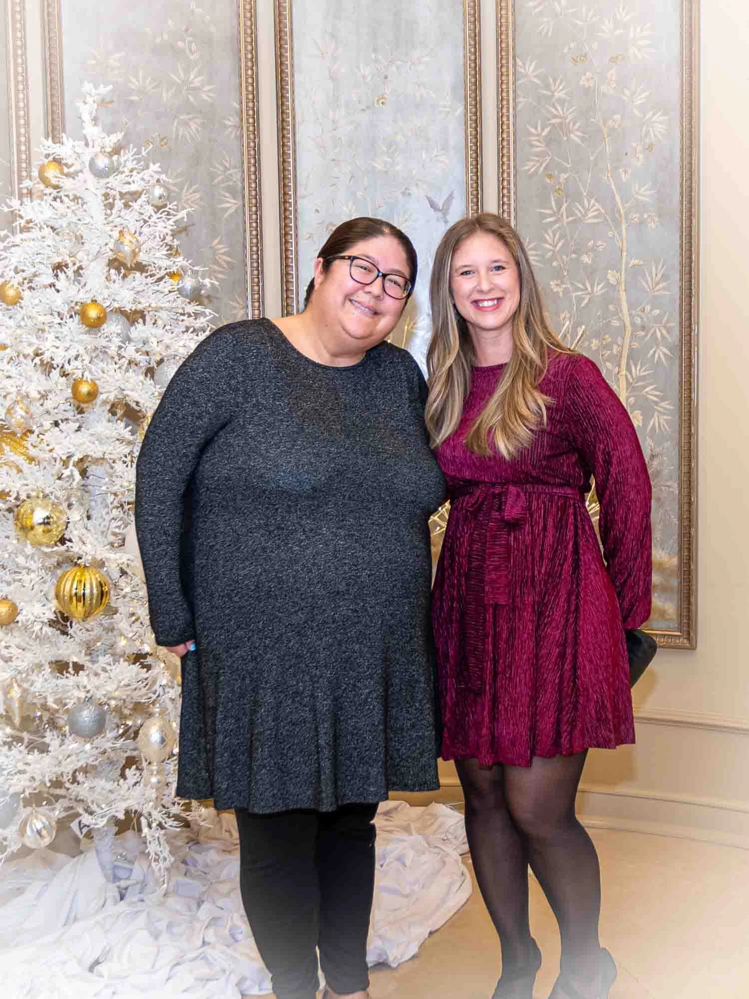 Two women standing in front of a decorated white Christmas tree, smiling, with an ornate wall in the background. One is wearing a gray dress and leggings, the other a red dress.