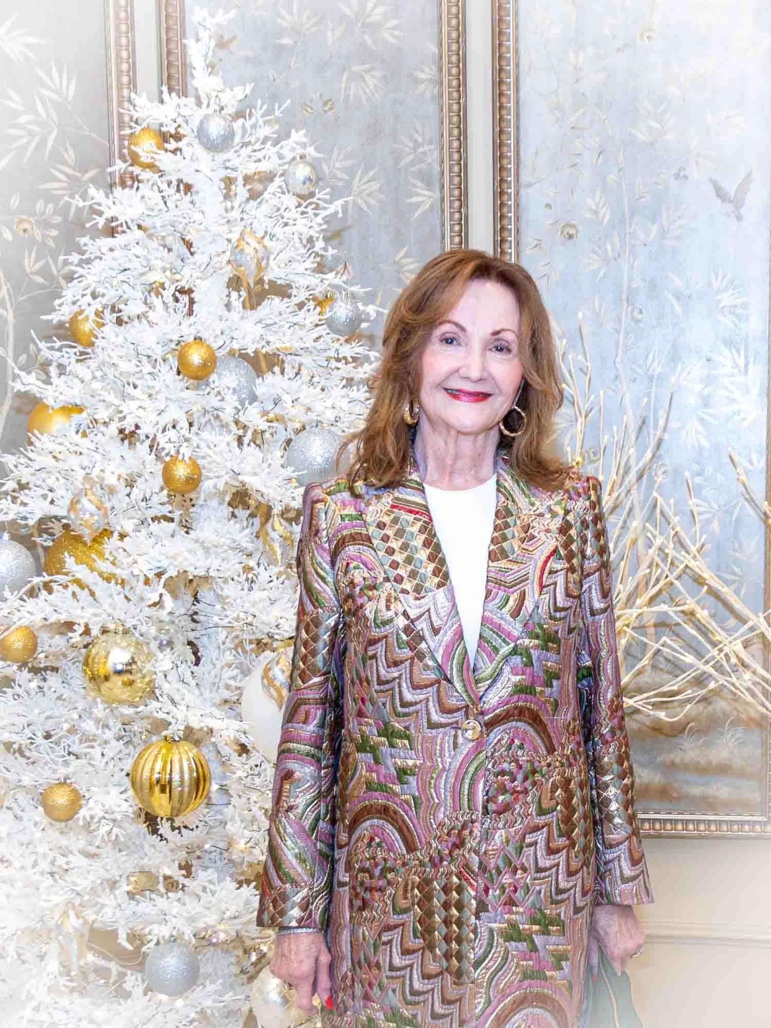 Woman in colorful patterned suit standing beside a white Christmas tree with gold and silver ornaments.