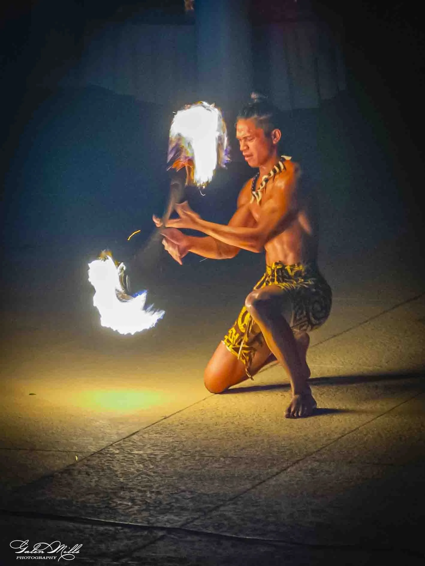 A male fire dancer performing at night using fire poi, kneeling with one knee on the ground, wearing traditional patterned shorts, and holding flaming poi that create bright, fiery trails.