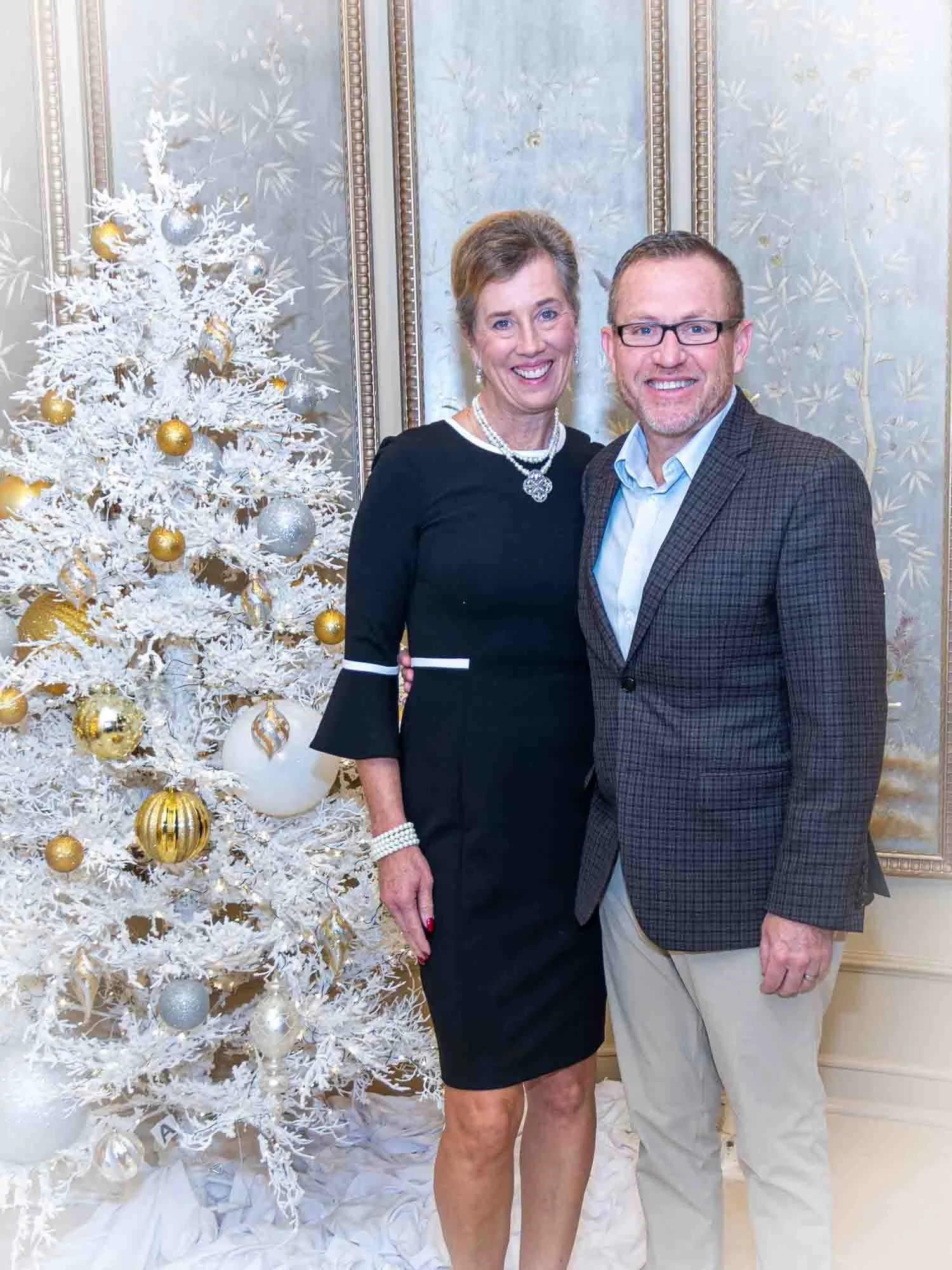 Two people posing in front of a decorated white Christmas tree with gold and silver ornaments, in a elegant room with ornate wallpaper.