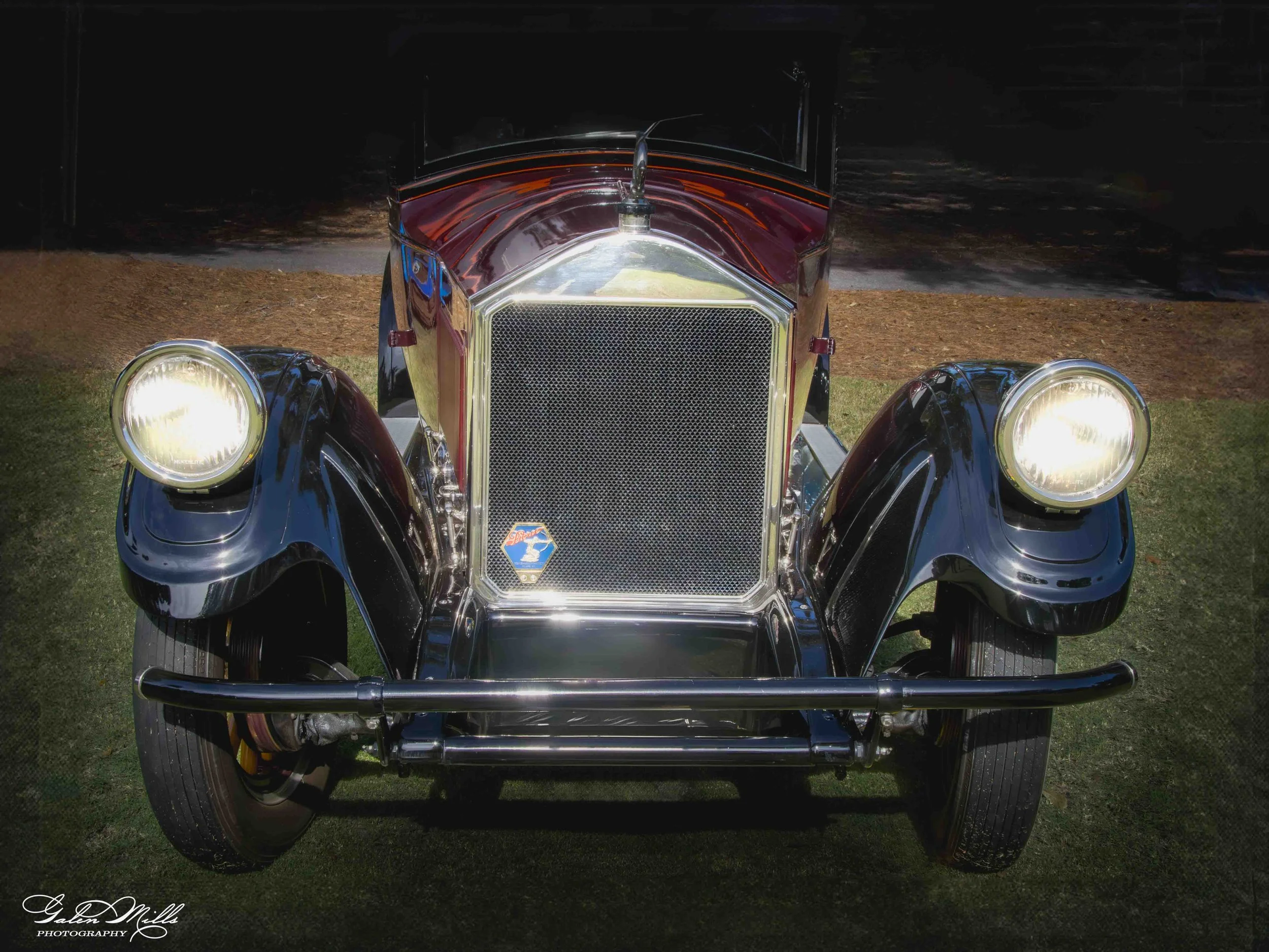Front view of a vintage black and chrome automobile with round headlights and a prominent grille, parked outdoors on grass with a dark background.