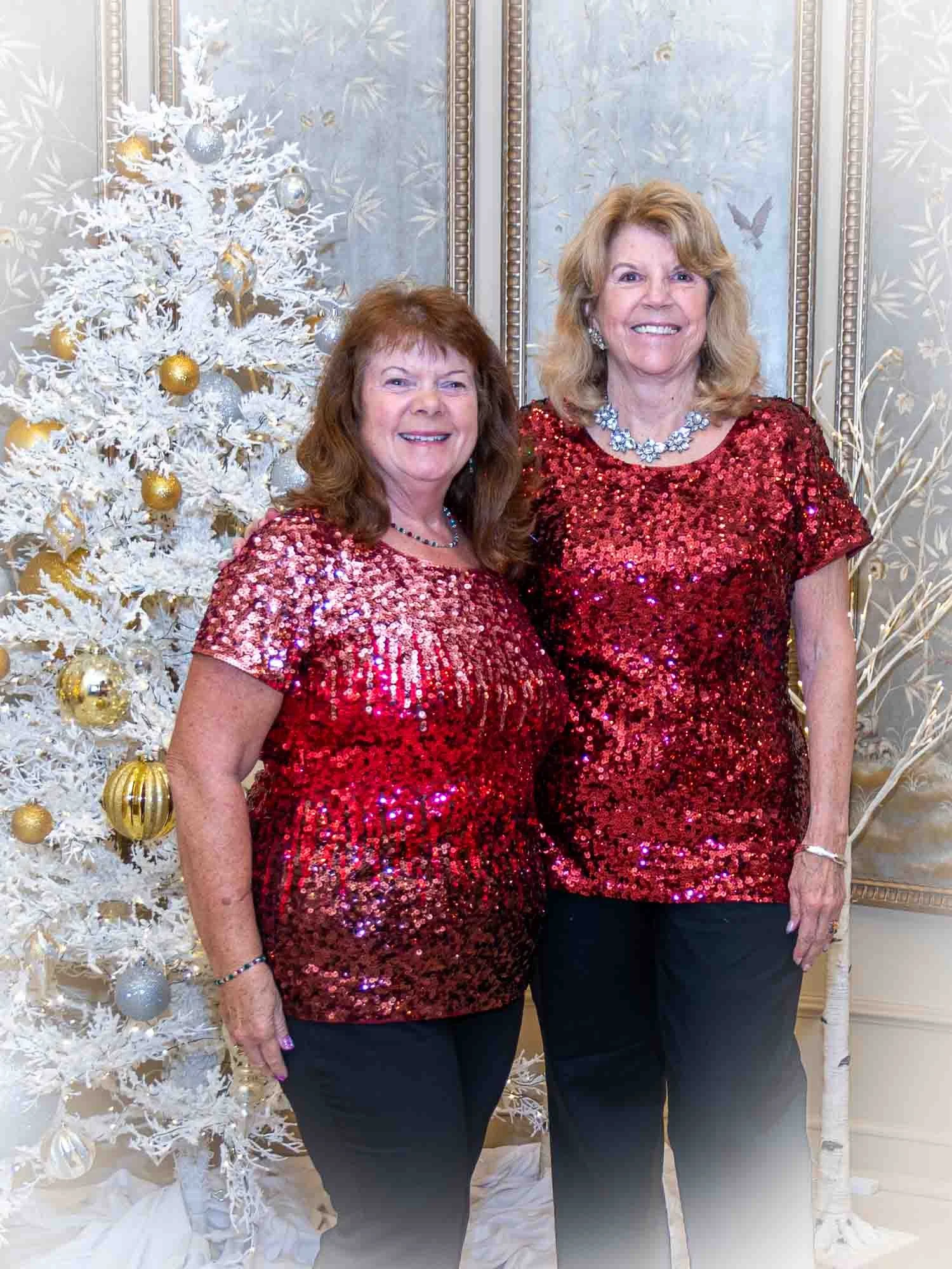 Two women wearing red sequined tops standing in front of a white Christmas tree with gold and silver ornaments.