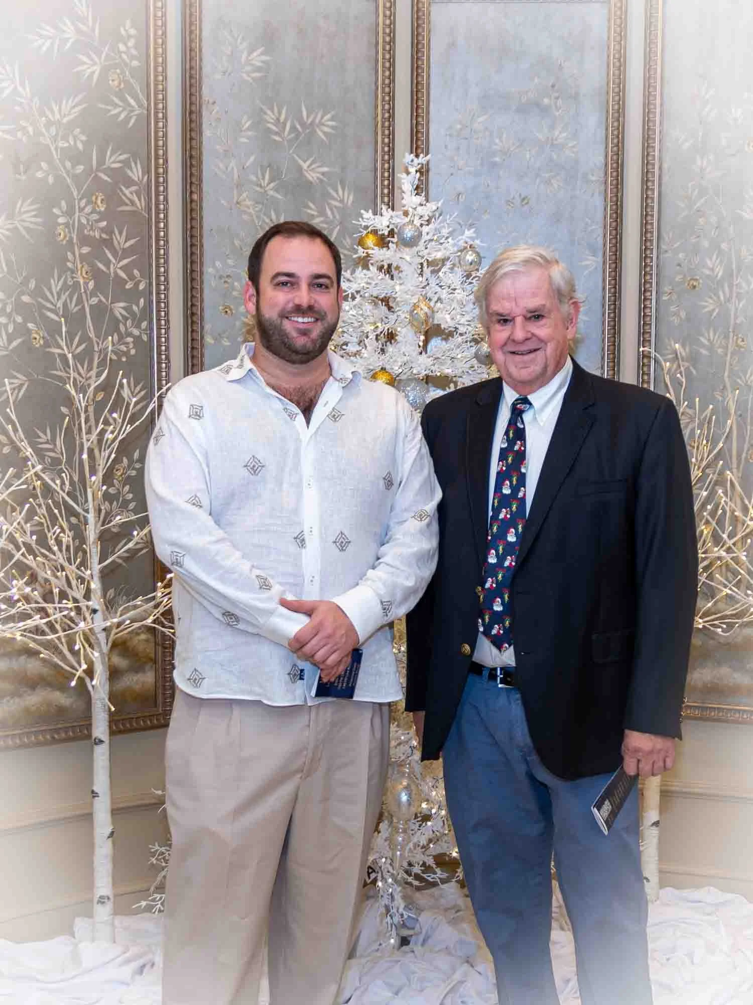 Two men posing together in front of white Christmas decorations, including a white tree with gold and silver ornaments, in a formal indoor setting.