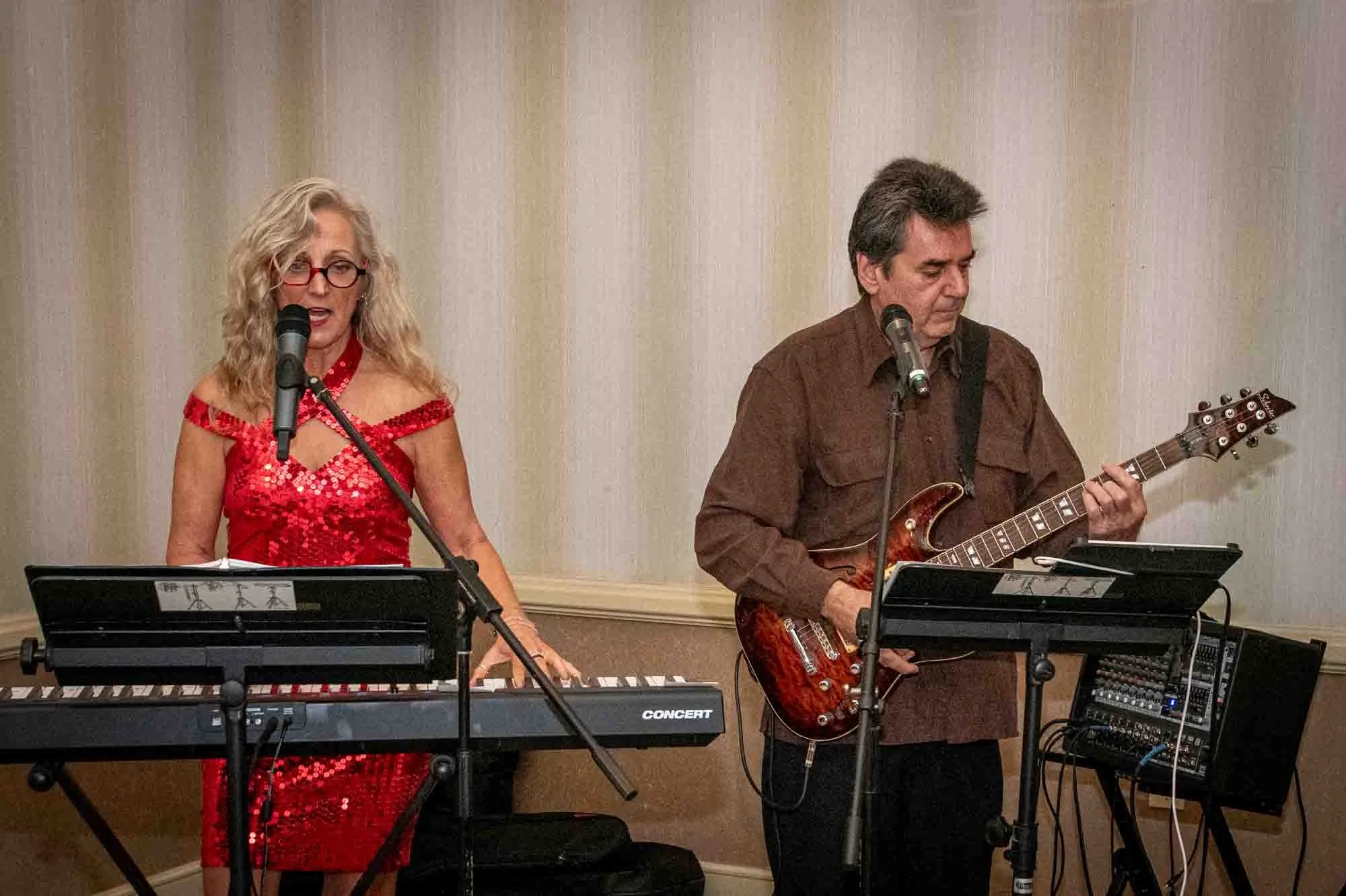 A woman in a red sequined dress playing a keyboard and a man playing an electric guitar, both singing into microphones.