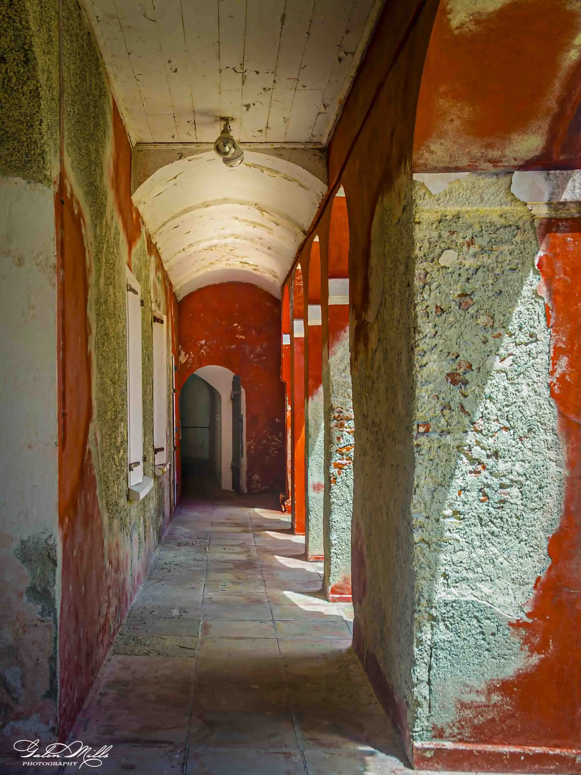 A long, narrow hallway with an arched ceiling. The walls are partially painted with a mix of red, green, and white colors, and some areas appear to be unfinished or in renovation. There is a single light fixture hanging from the ceiling at the front.