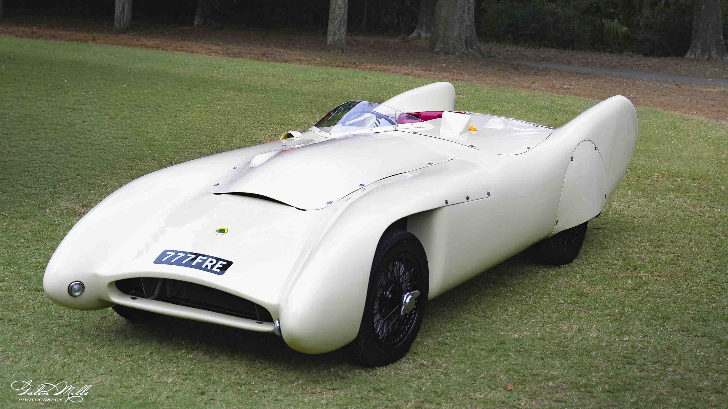 A vintage white race car with a British license plate '777FRE' parked on a grassy field with trees in the background.