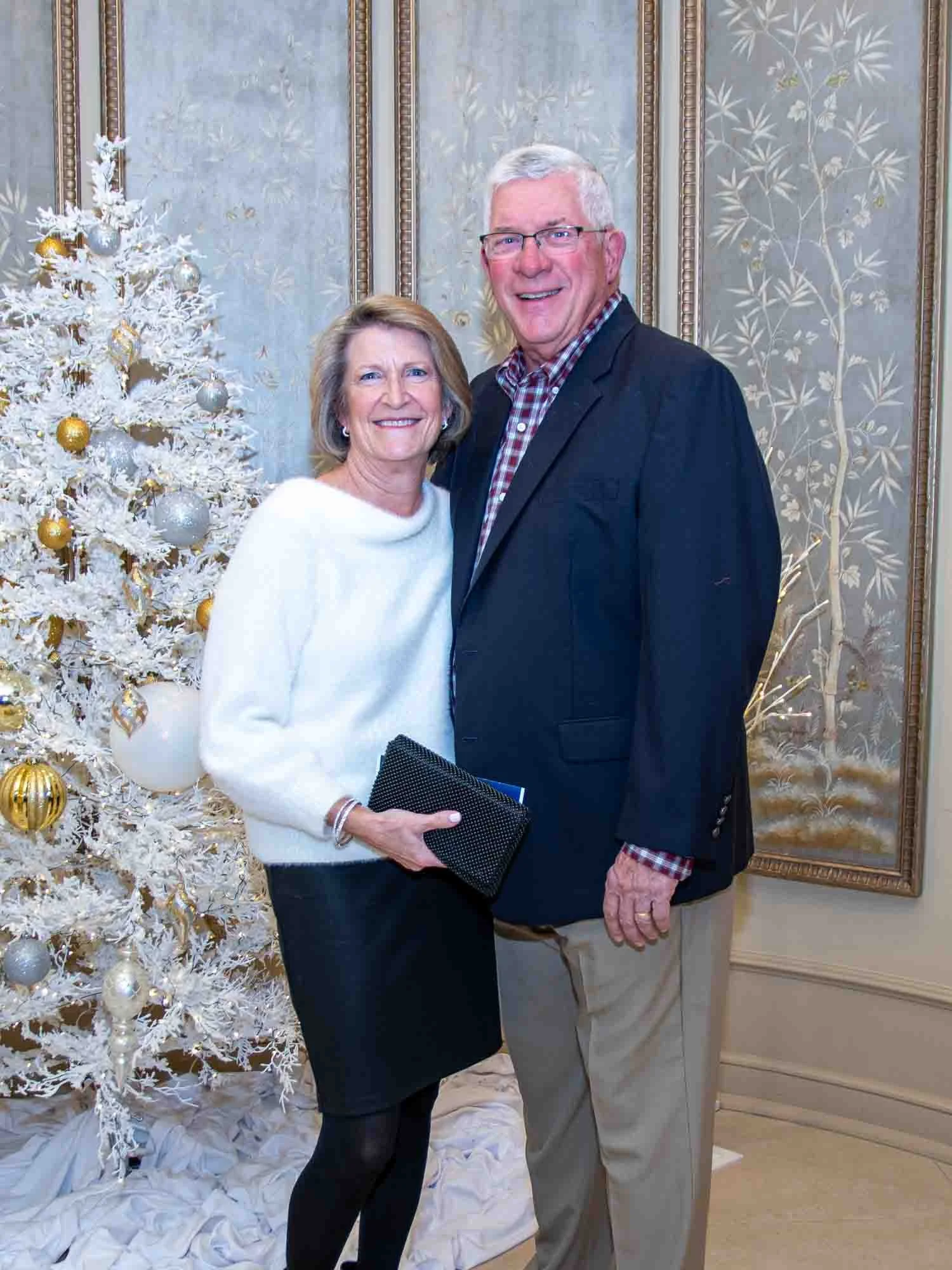 A couple posing in front of a decorated white Christmas tree with gold and silver ornaments, standing in an elegantly designed interior with patterned wallpaper.