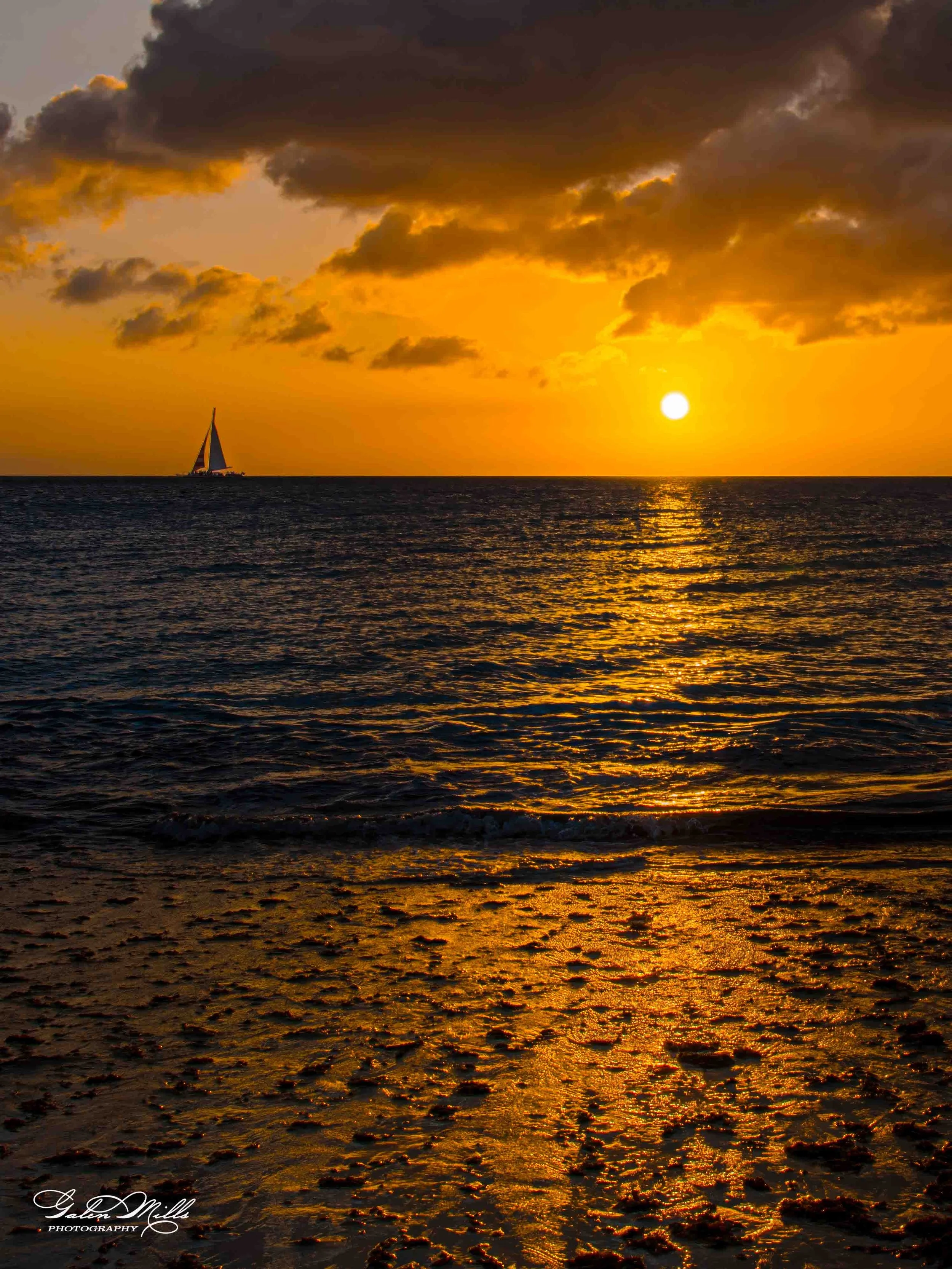 Sunset over the ocean with dark clouds in the sky and a sailboat sailing on the horizon. The reflection of the sun creates a golden path on the water and beach.
