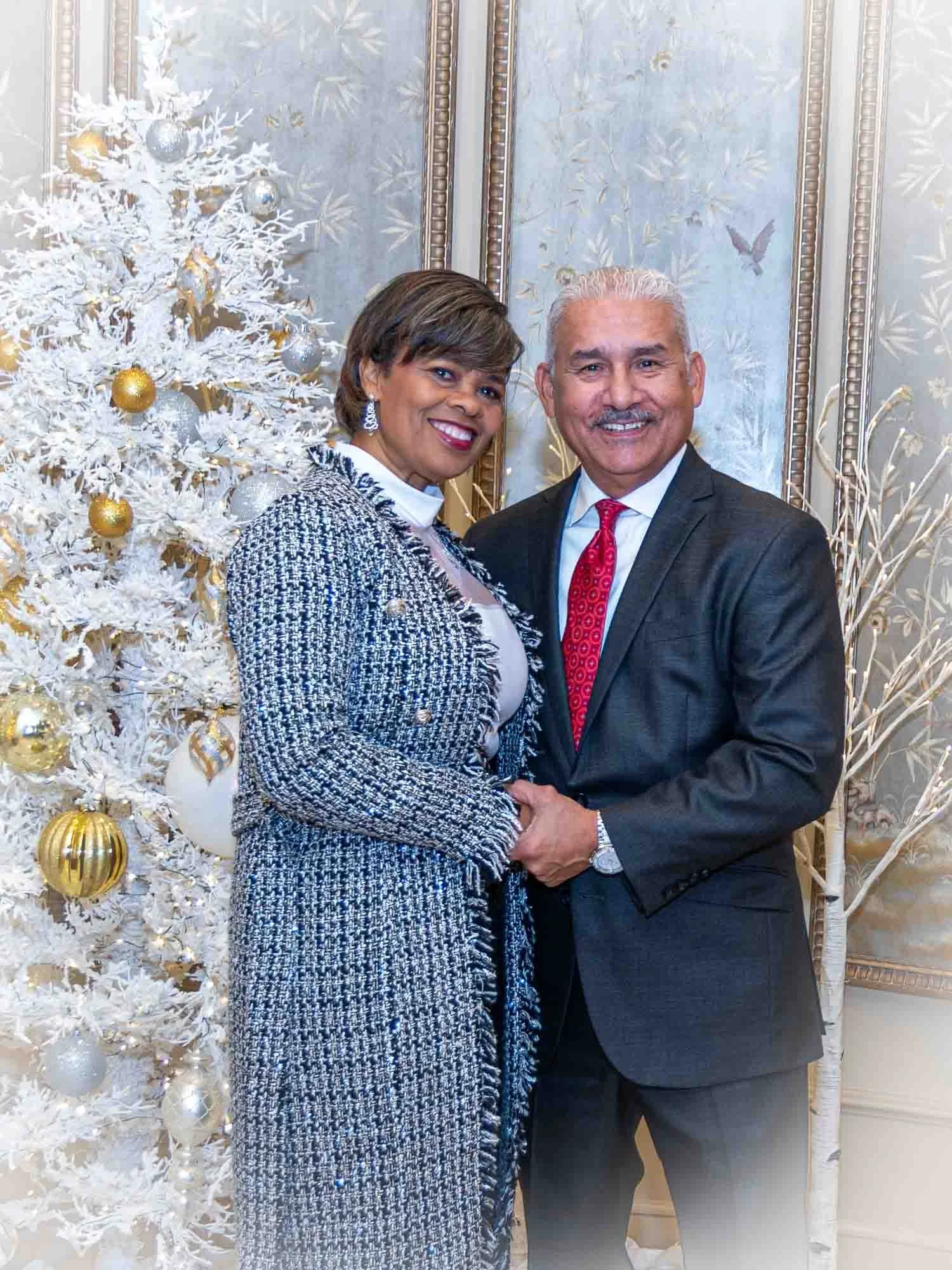 A smiling couple standing next to a white Christmas tree decorated with silver and gold ornaments, in an elegant room with ornate wallpaper.