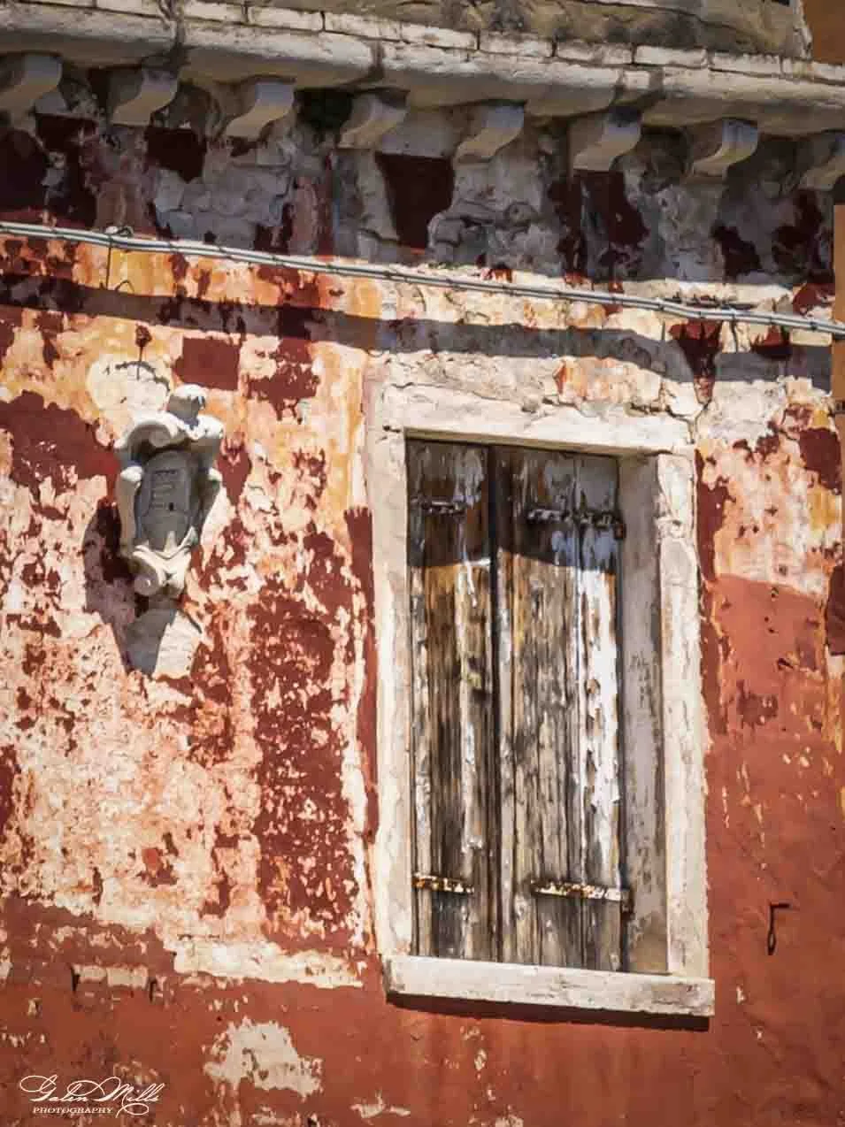 An old building with a weathered, red-painted exterior wall, a rustic wooden window with closed shutters, and an ornate wall-mounted lamp fixture.