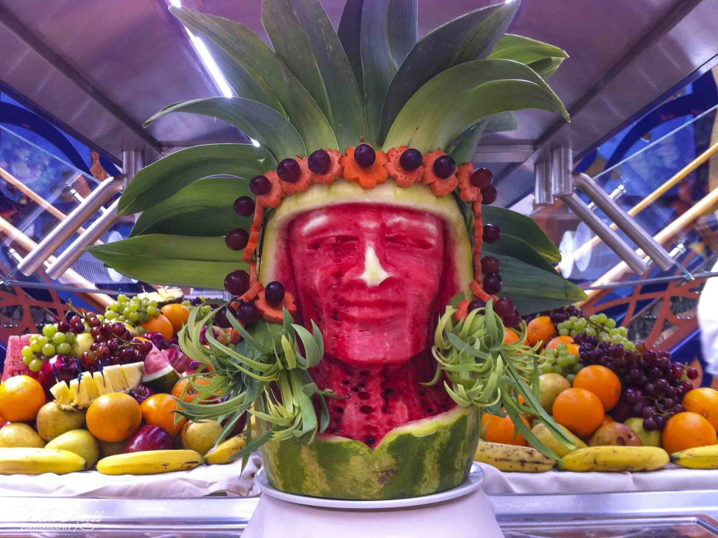 A decorative display featuring carved watermelon at the center, surrounded by various fruits including oranges, bananas, apples, grapes, and other colorful fruits, with tropical leaves on top.