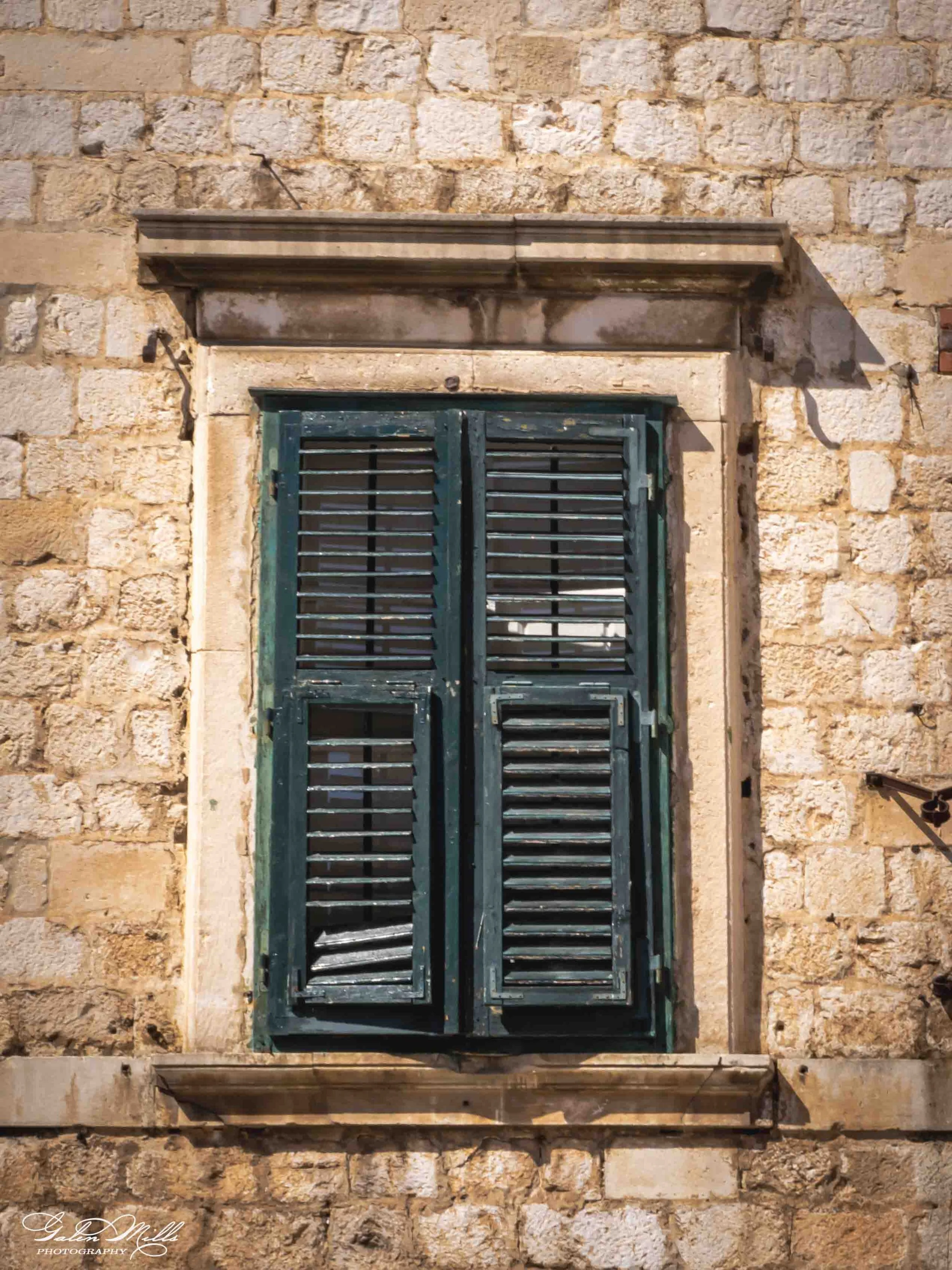 A window with green shutters mounted on a stone wall. The shutters are closed and the wall is built with rough, light-colored stones.