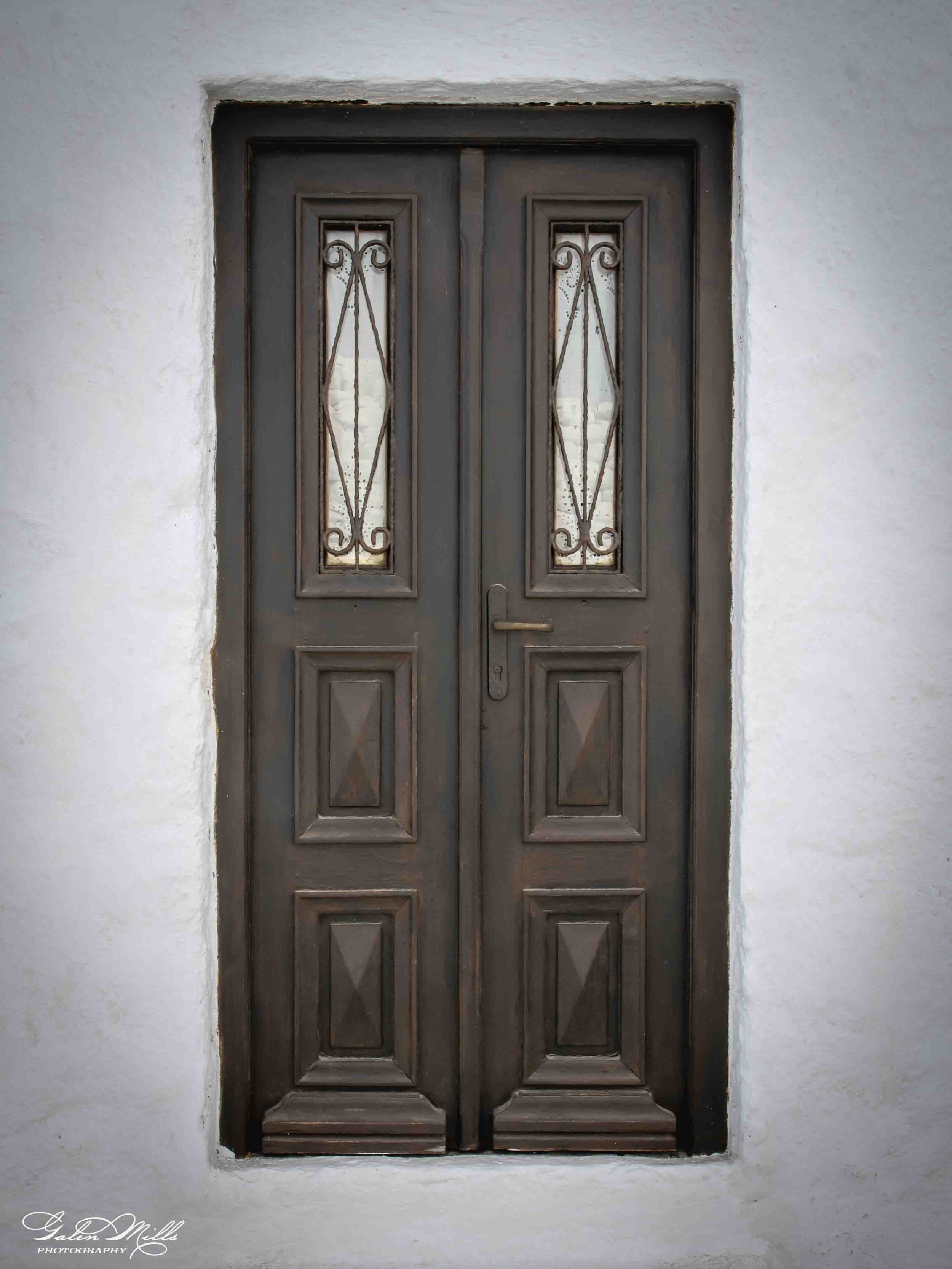 A closed, brown double door with decorative metal grills and glass panels, set in a white wall with a rectangular opening.