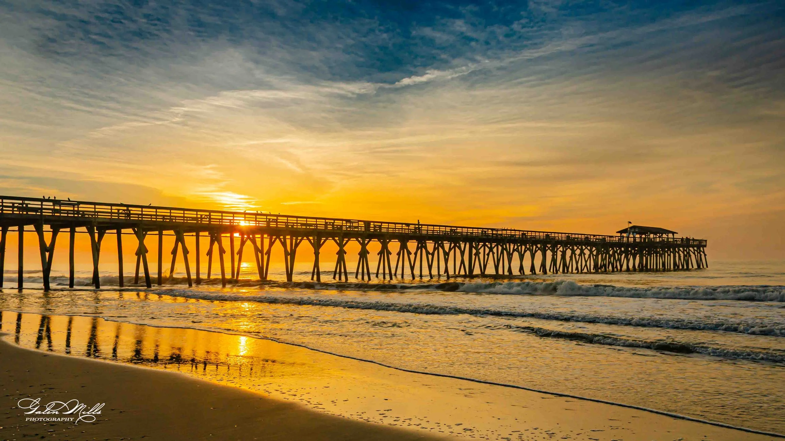Sunset over the ocean with a wooden pier extending into the water, some birds sitting on the pier, and gentle waves on the shore.