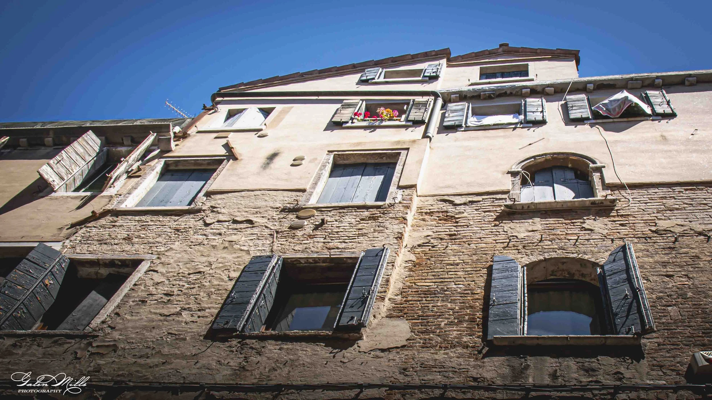 An upward view of an old building with broken and open black shutters, some with flowers, and a weathered brick and stucco facade against a clear blue sky.