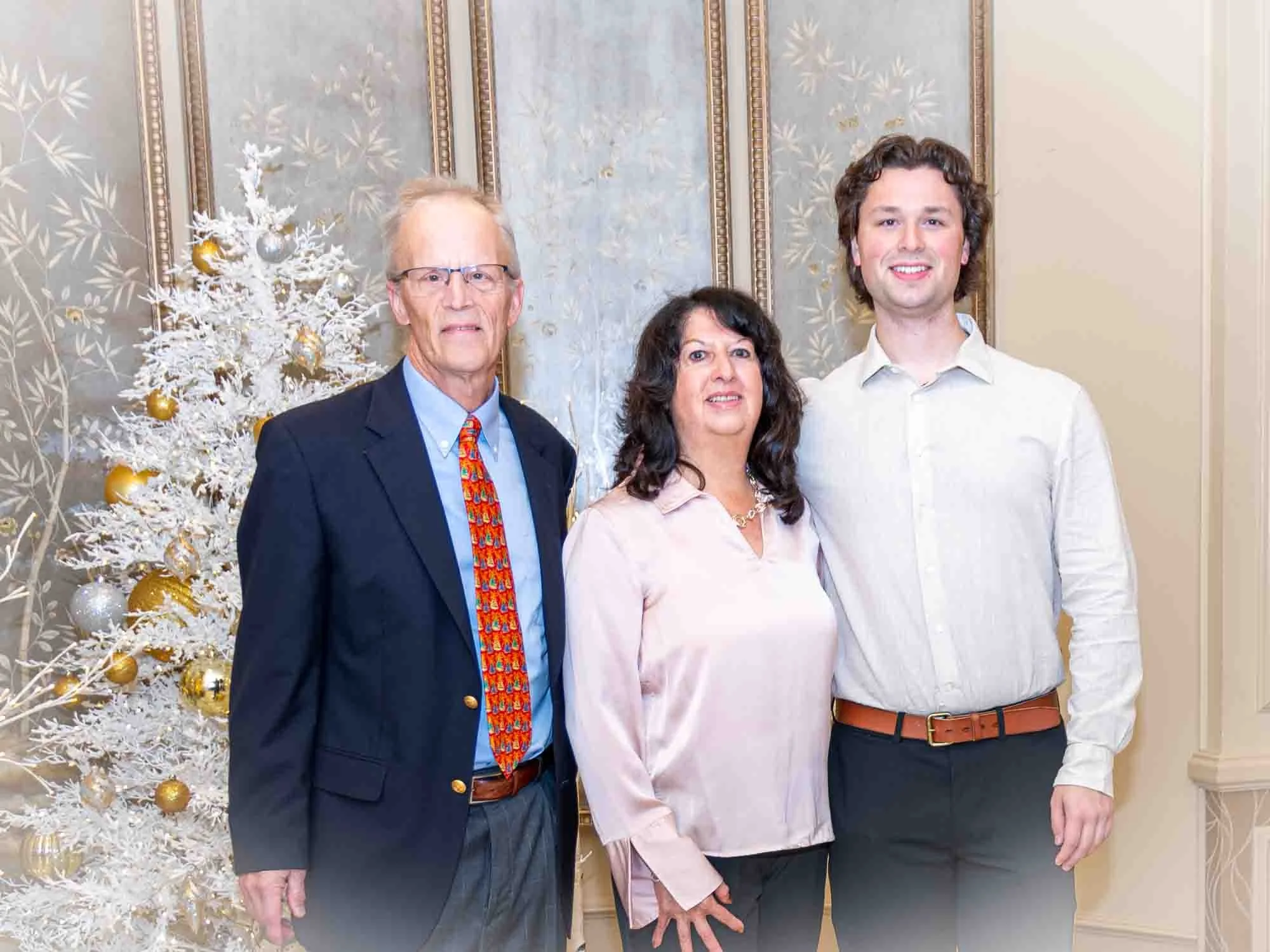 Three people standing indoors, a decorated white Christmas tree in the background, dressed in formal attire.