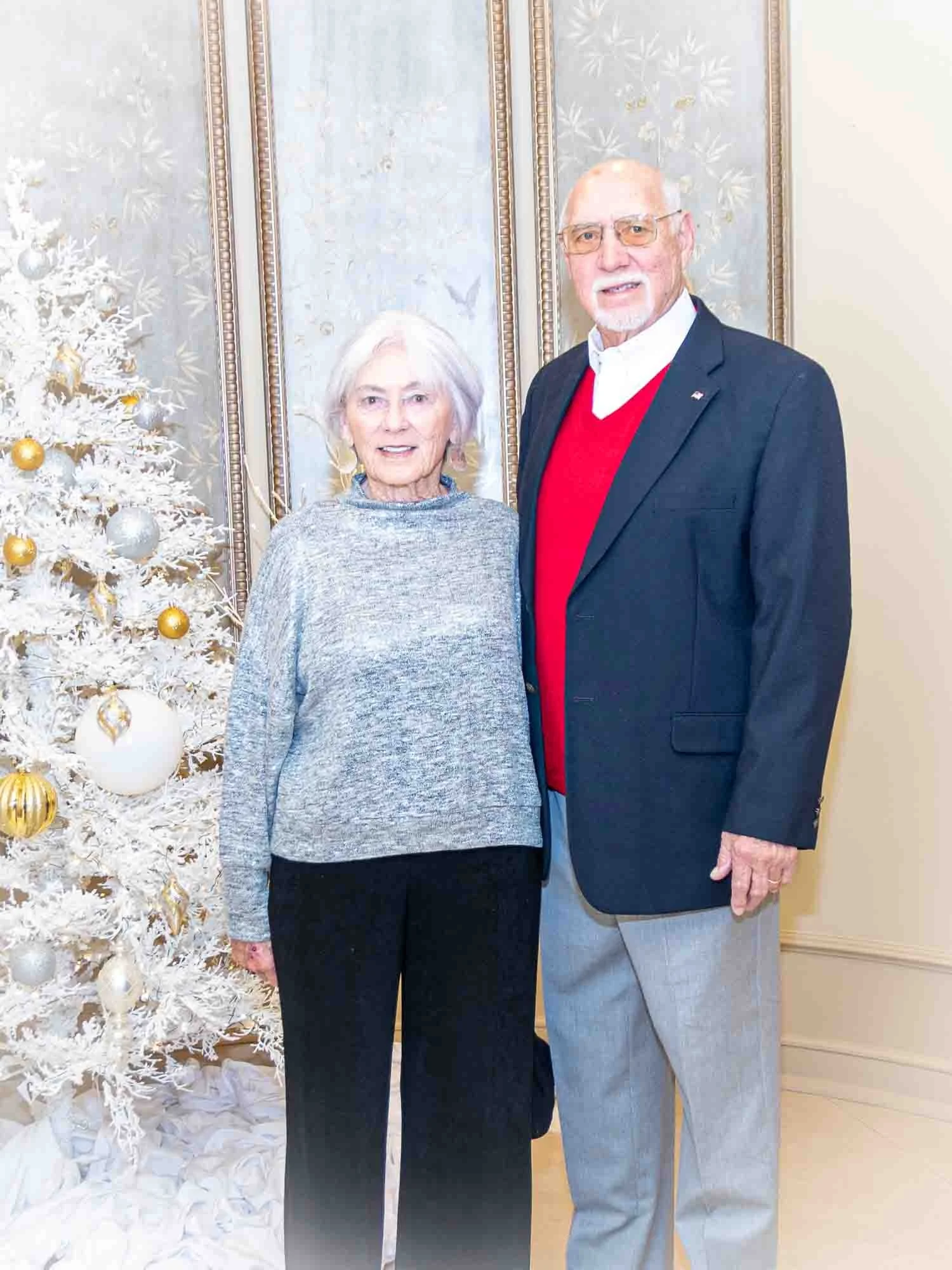 An elderly couple stands beside a white Christmas tree decorated with gold and silver ornaments. The woman is wearing a gray sweater, and the man is wearing a dark blazer with a red sweater underneath. They are smiling in a warmly lit room.