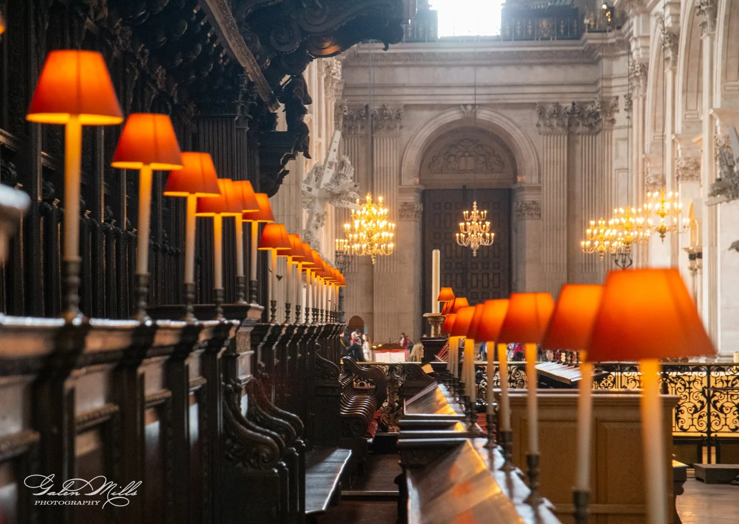 Interior of a cathedral featuring ornate wooden pews, orange-shaded lamps, chandeliers, and detailed architectural elements.