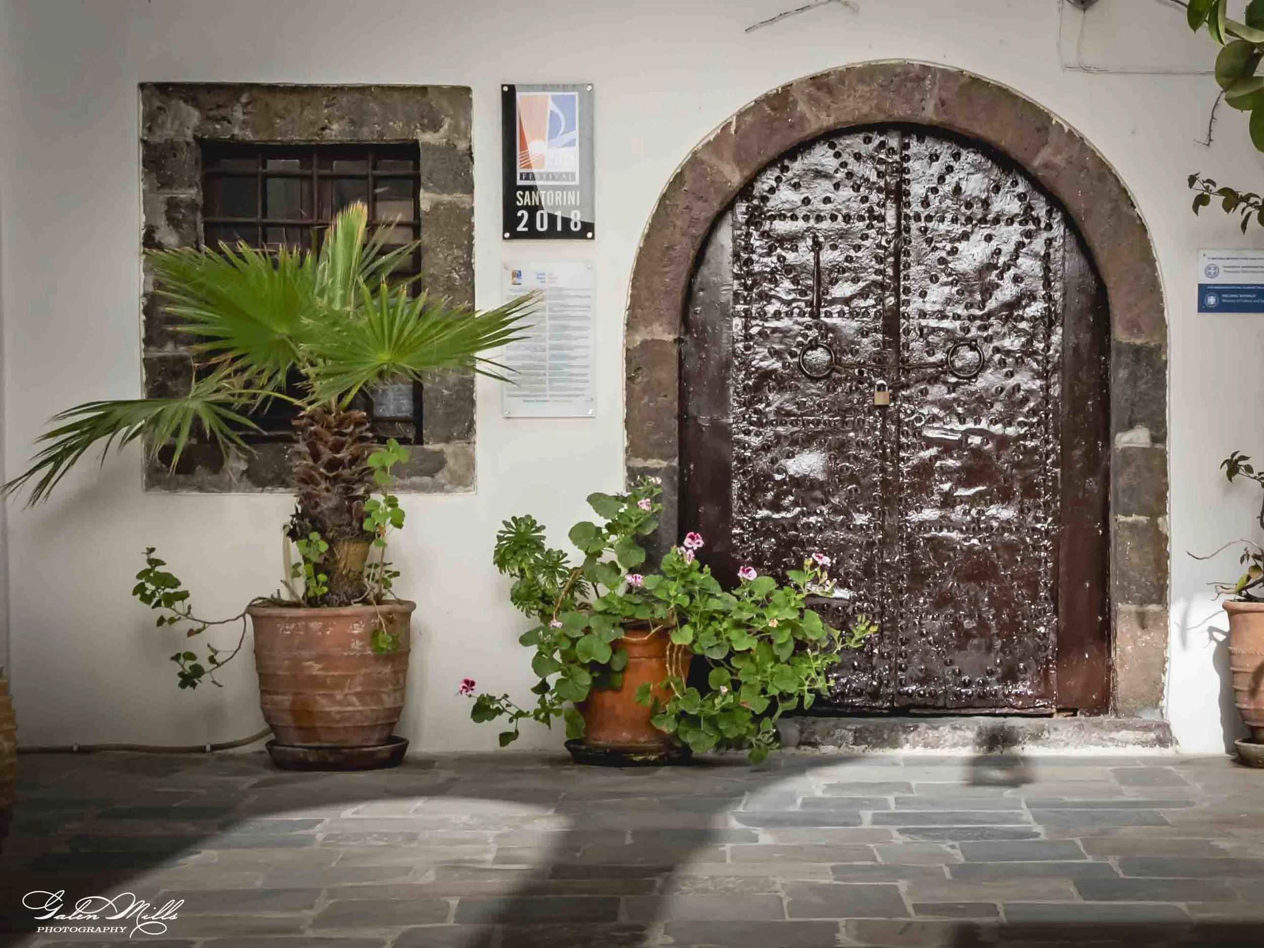 A rustic, arched, metal door with rivets and a lock, surrounded by white walls. There are potted plants with green leaves and pink flowers on either side of the door. A sign reads 'Santorini 2018,' and there are small windows and wall decorations nea