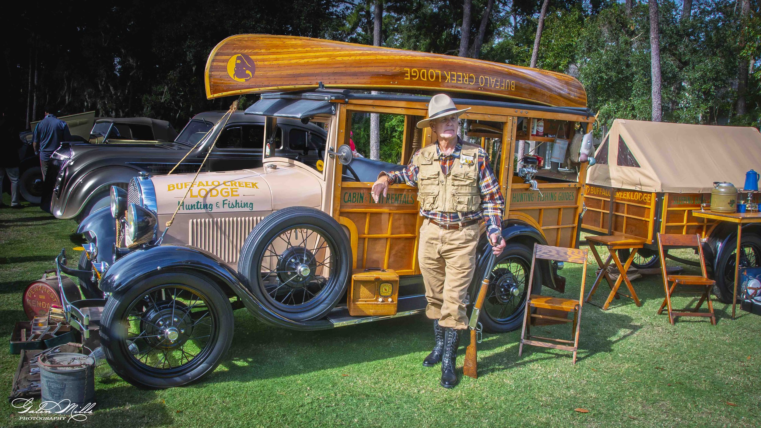 Vintage truck with wooden canoe on top parked outdoors, man in safari hat and khaki vest standing beside, green trees in background.