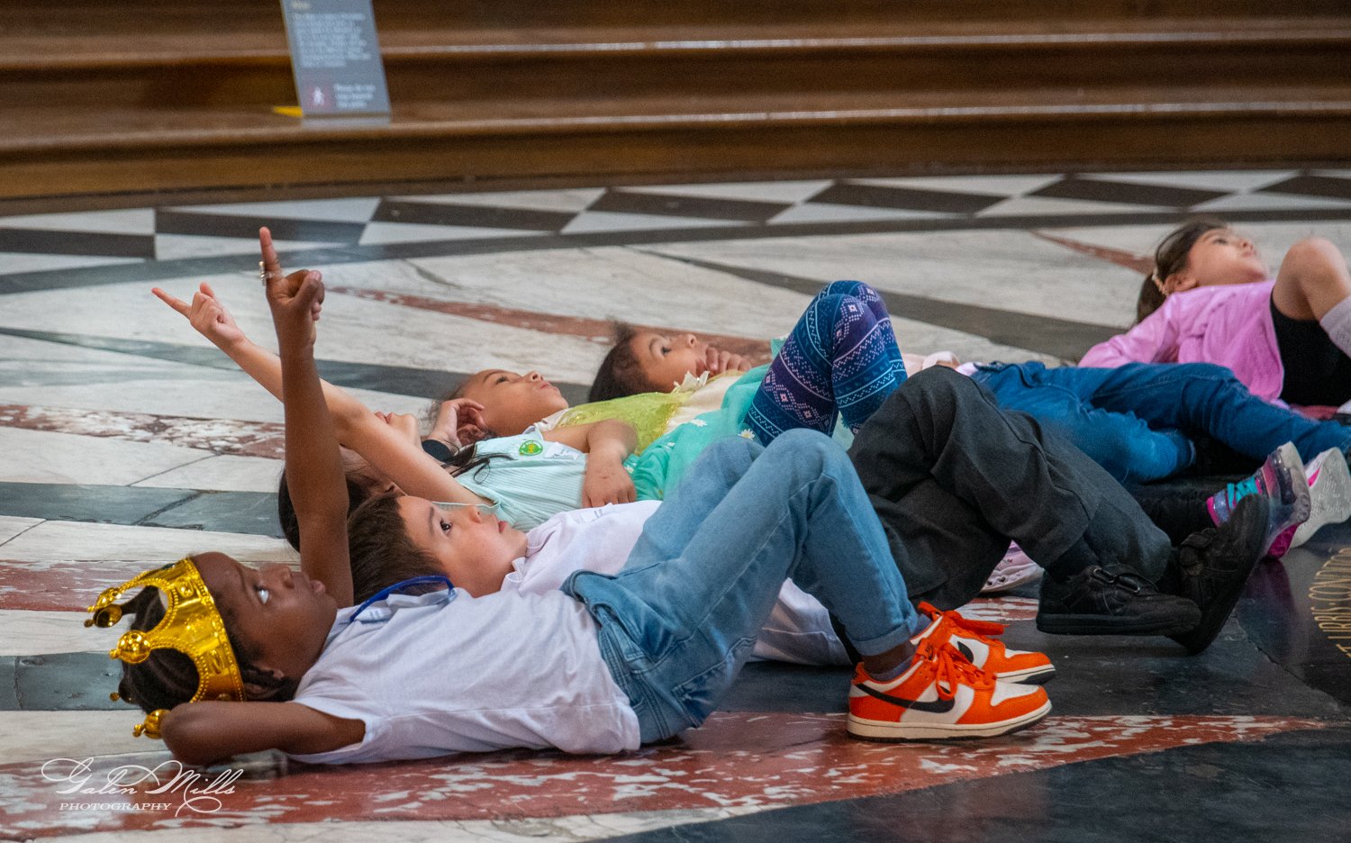 Children lying on a patterned floor, looking and pointing upwards.