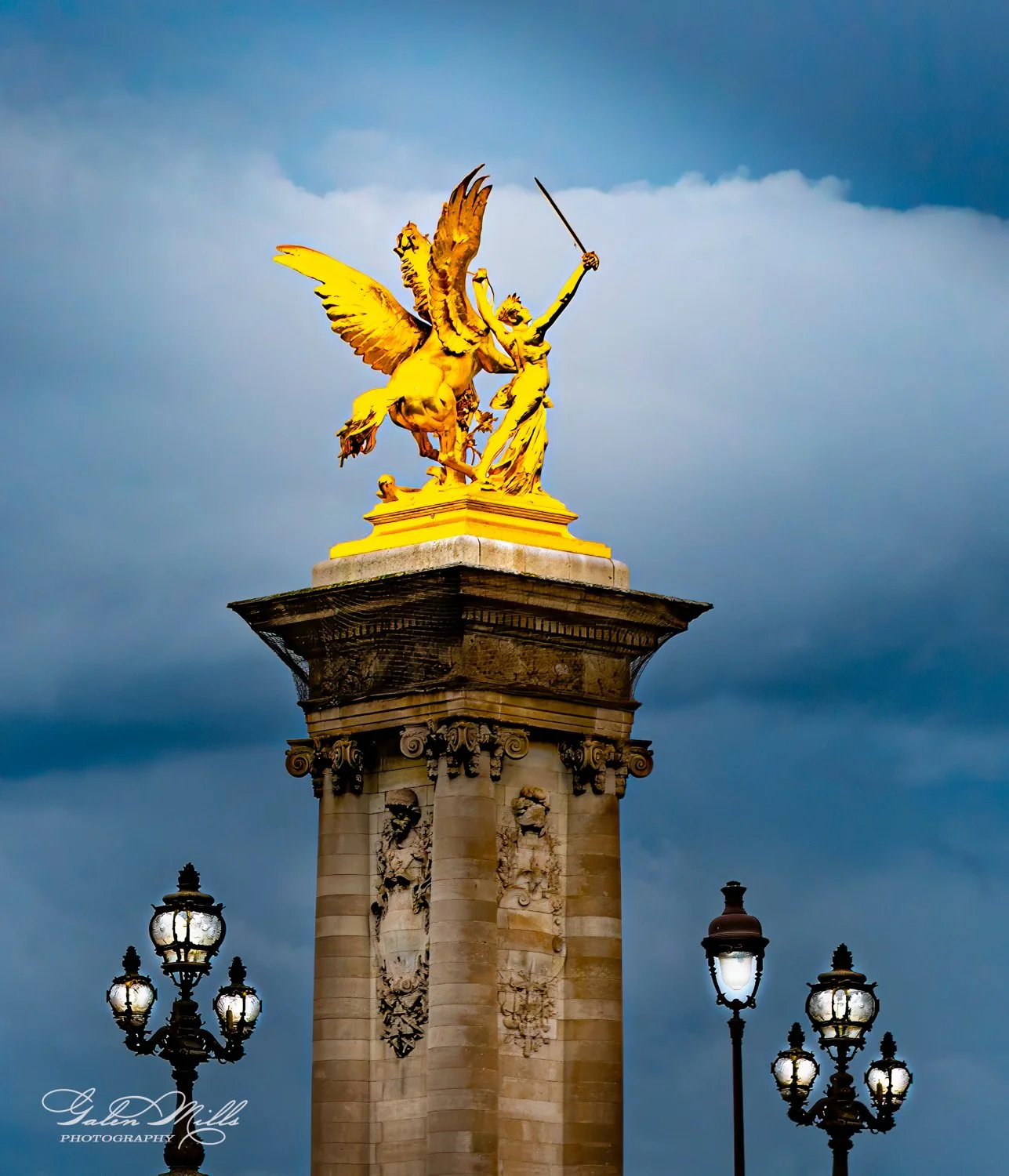 Golden statue of a winged horse and a warrior on a pillar, with ornate street lamps, against a cloudy sky.