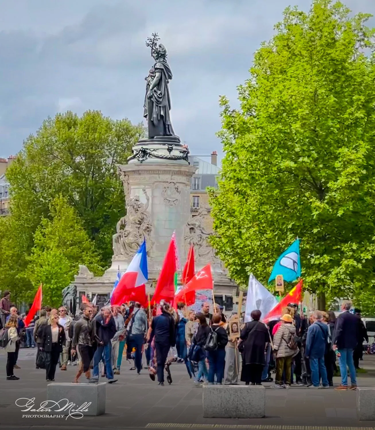 A group of people gathered in front of a large statue, holding various colorful flags, including red and a French flag, surrounded by trees.