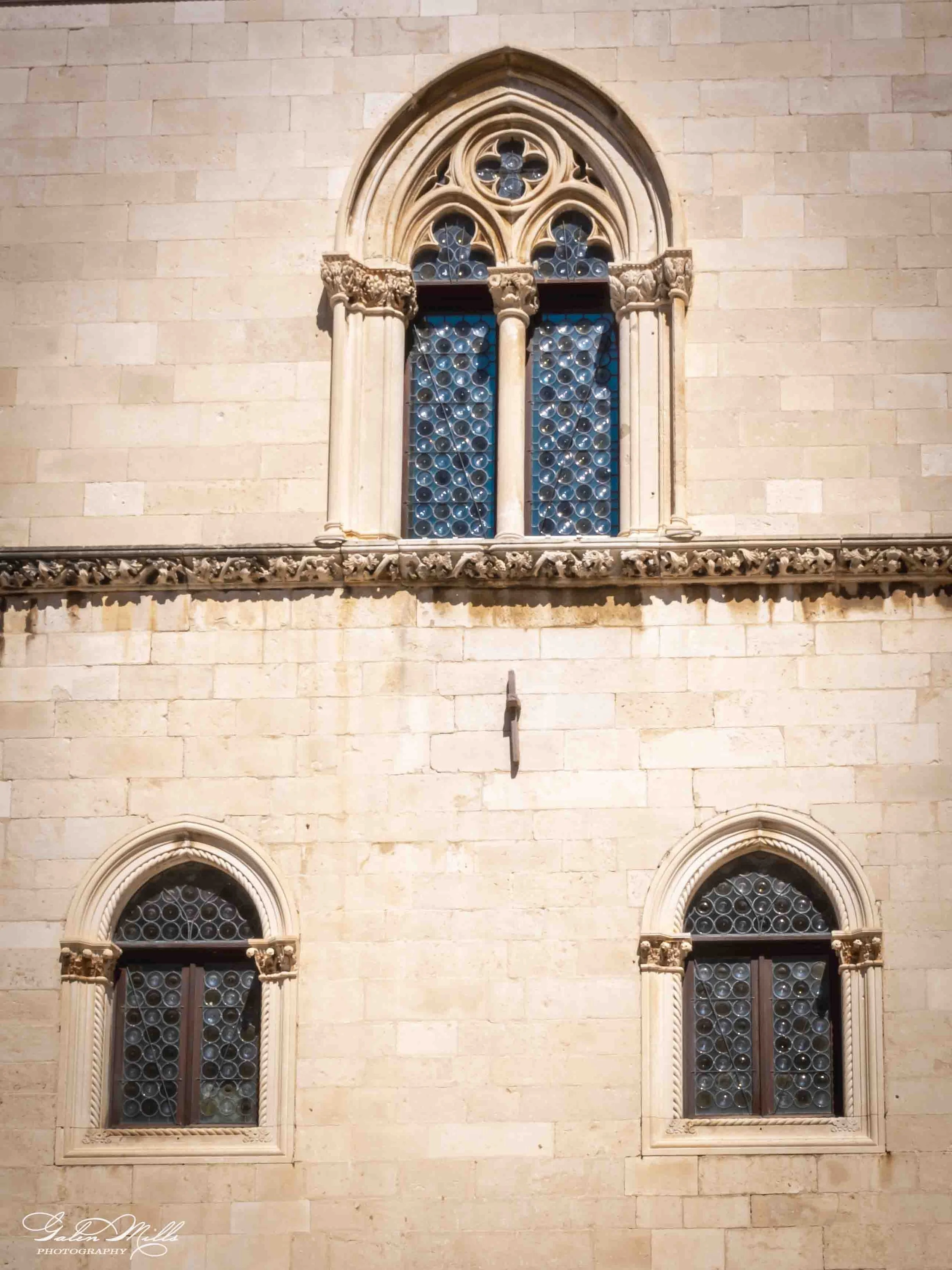 The image shows a close-up view of a stone building's facade with three decorative arched windows with intricate trim and stained glass. The building's wall is made of light-colored stone blocks.