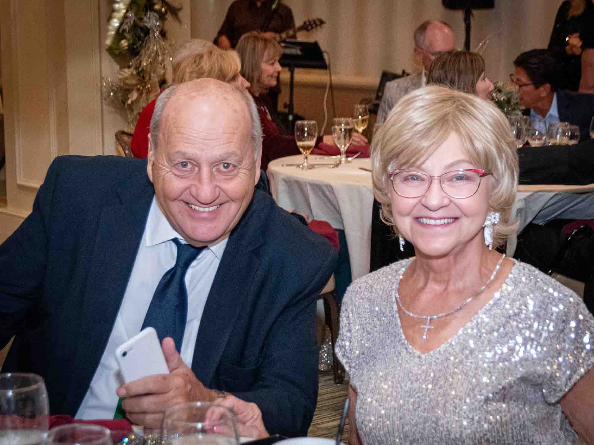 A smiling older couple seated at an event, with the man wearing a dark suit and tie and holding a smartphone, and the woman in a glittery top and glasses. They are surrounded by tables set with glasses and decor in a festive indoor setting.