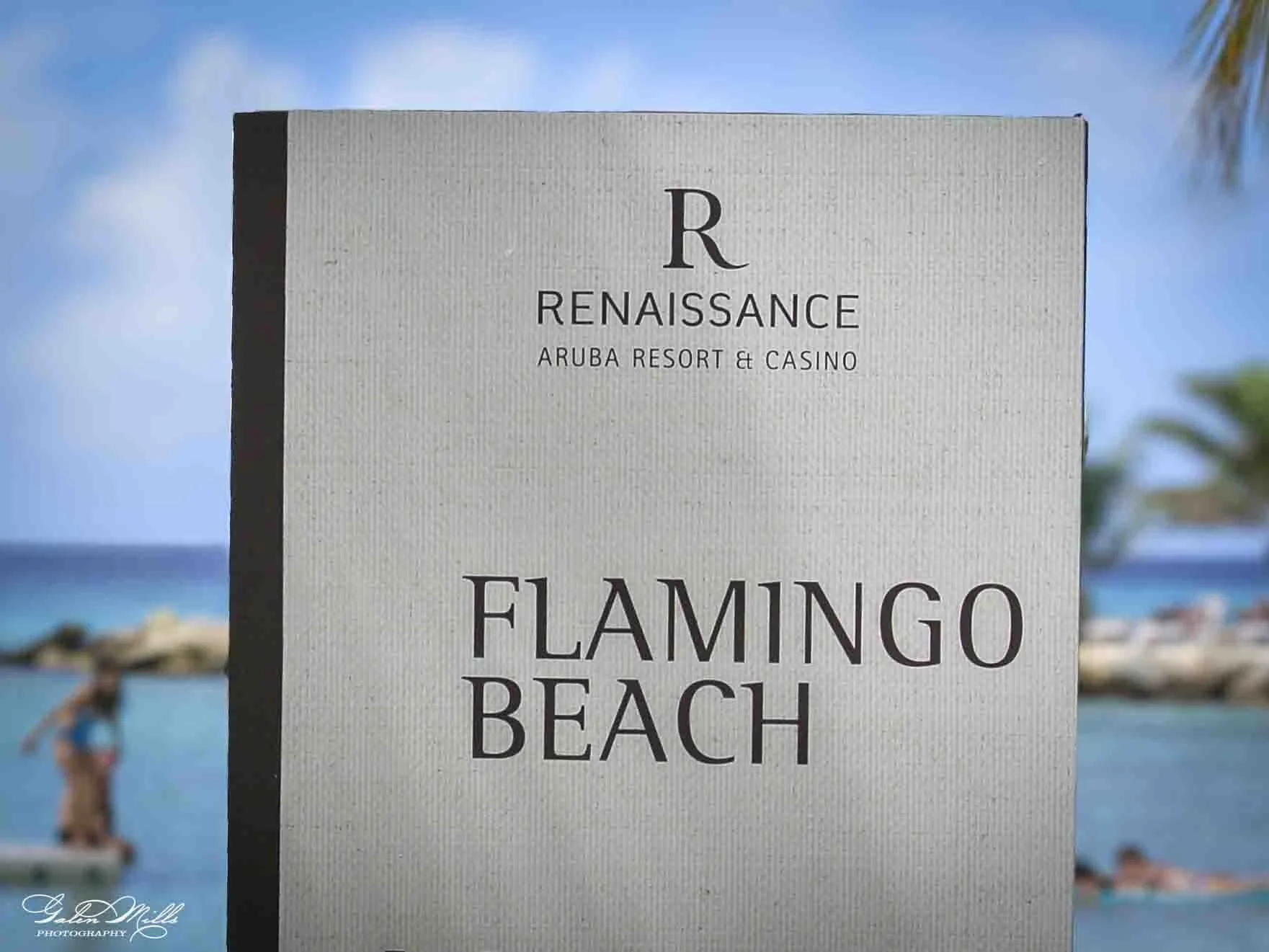 A sign reading 'Renaissance Aruba Resort & Casino' with the words 'Flamingo Beach' below. In the background, there is a woman taking a photo and blue sky with some clouds, beach, and palm trees.