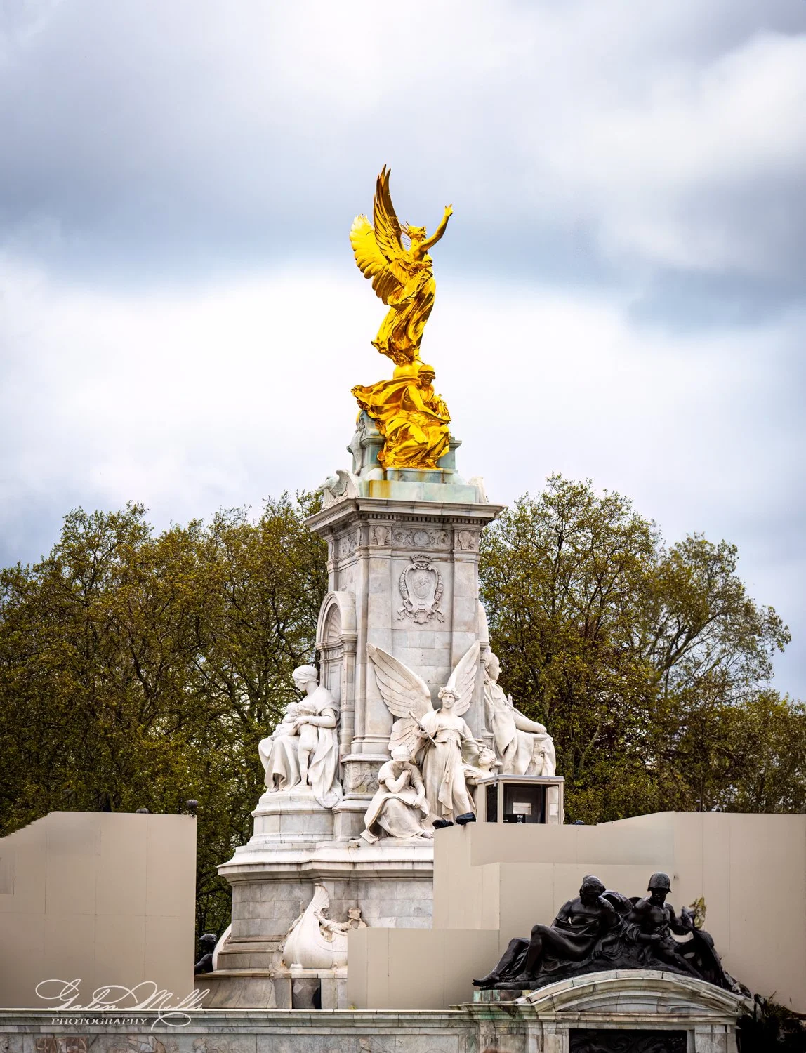 The Victoria Memorial in London, featuring a golden statue of Winged Victory atop a white stone base with figures and a cloudy sky backdrop.