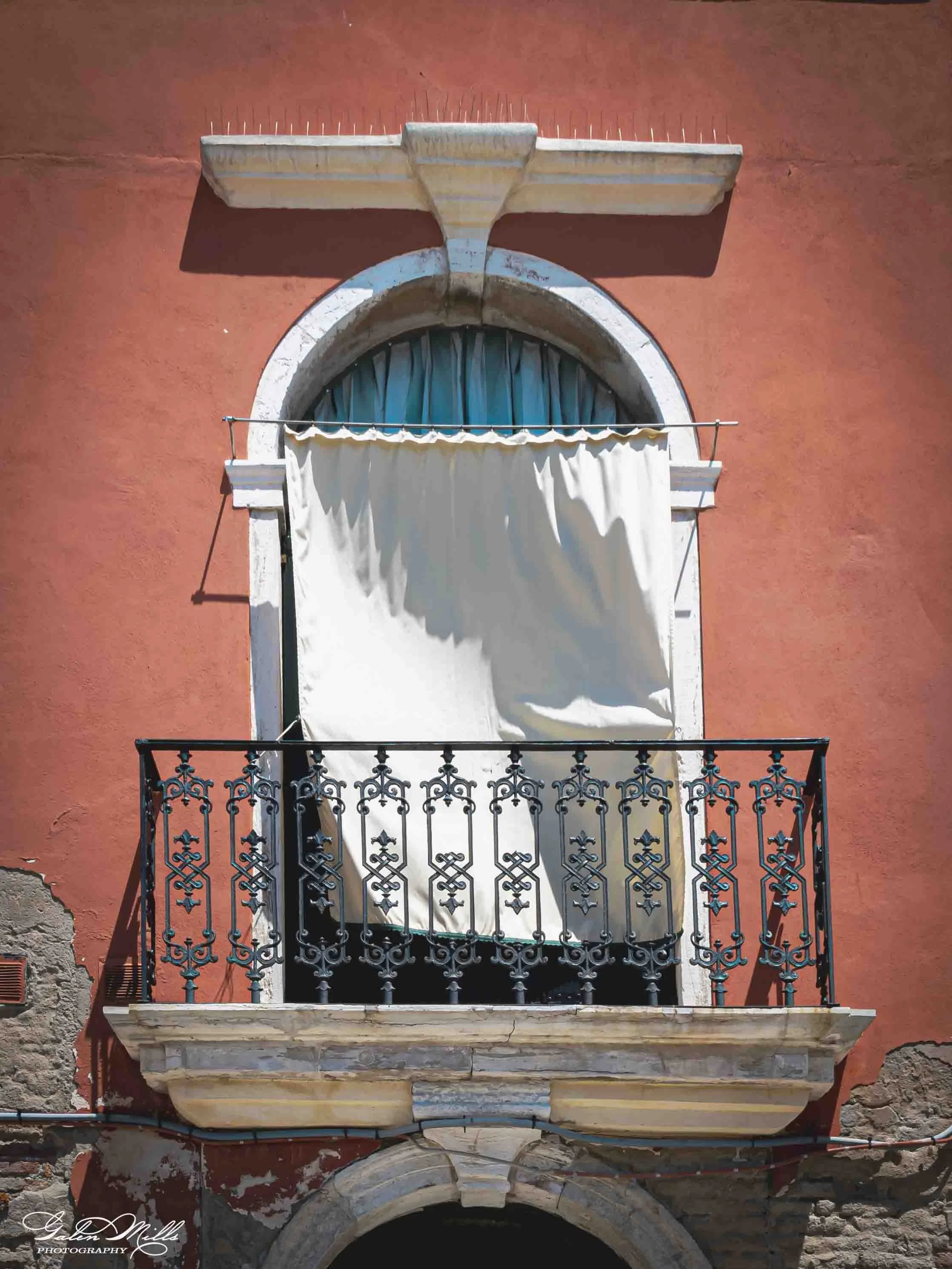 A building with a red wall, an arched window with a blue curtain, a small balcony with black ornate wrought iron railing, and a white fabric hanging below the window. The wall shows signs of wear and peeling paint.